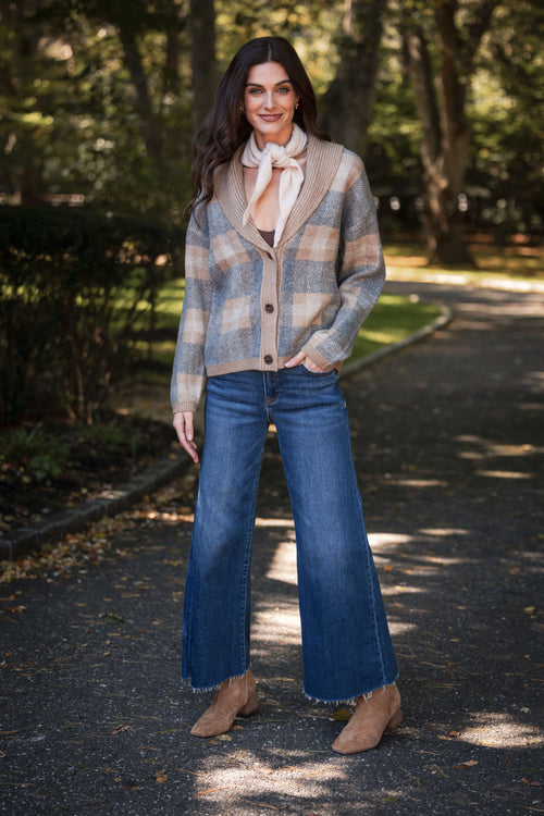 A woman stands outdoors wearing wide-leg blue jeans with frayed raw hems, a plaid cardigan, a cream neck scarf, and tan suede booties.