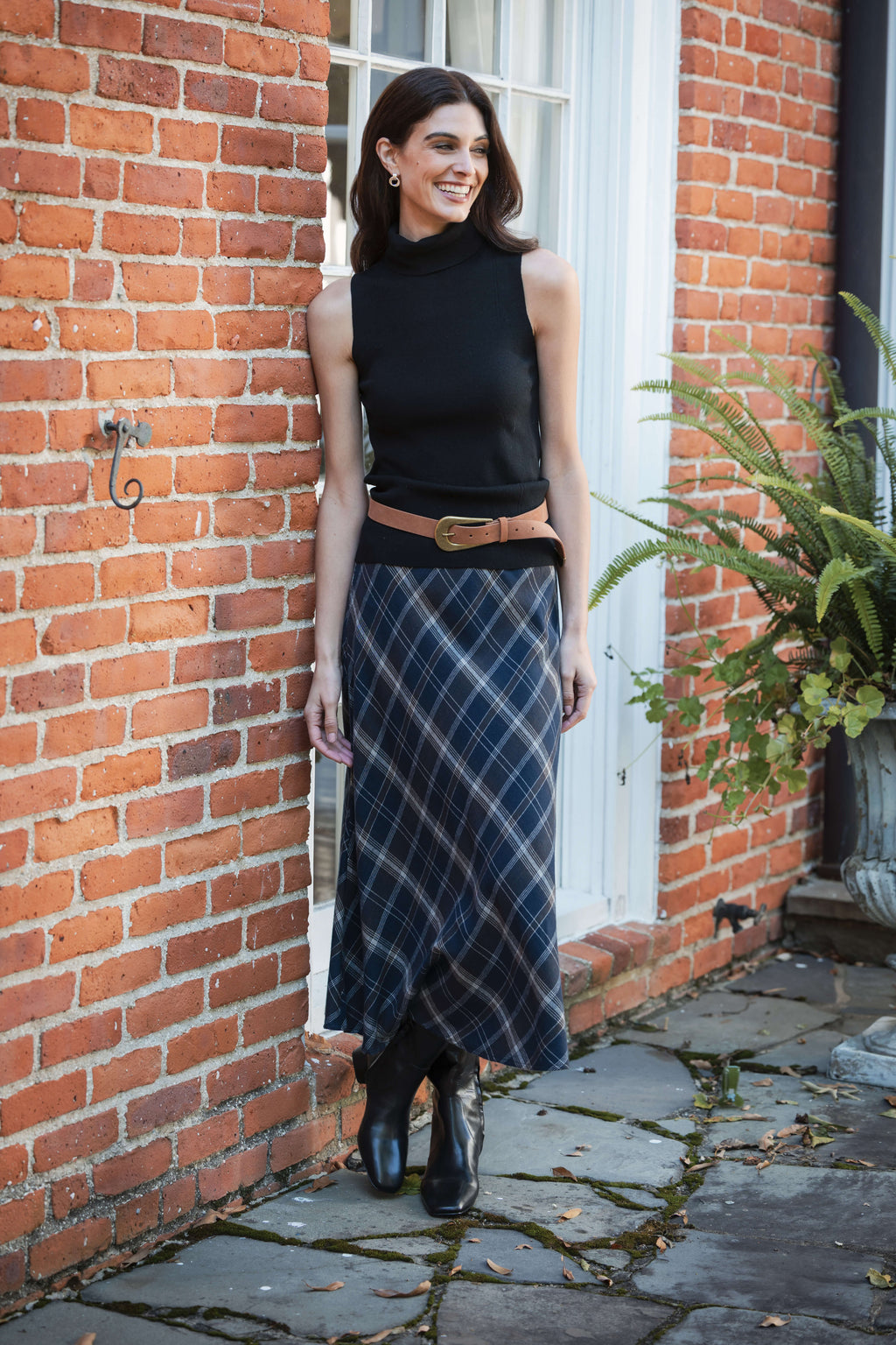 Black sleeveless mockneck top with cropped length worn with plaid skirt and tan belt.