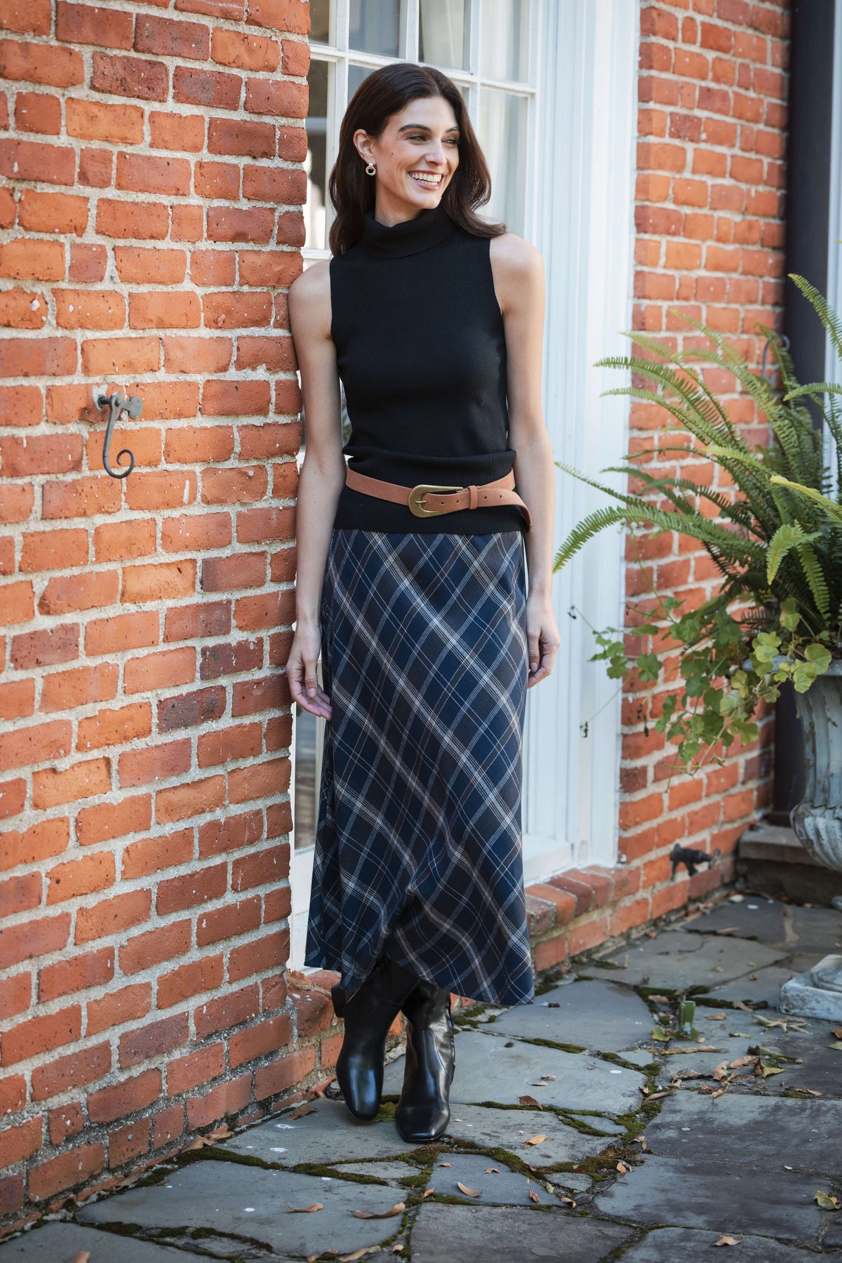 Black sleeveless mockneck top with cropped length worn with plaid skirt and tan belt.