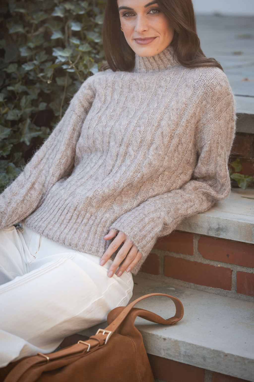 Beige cable knit turtleneck sweater on model wearing white pants, seated outdoors by water.