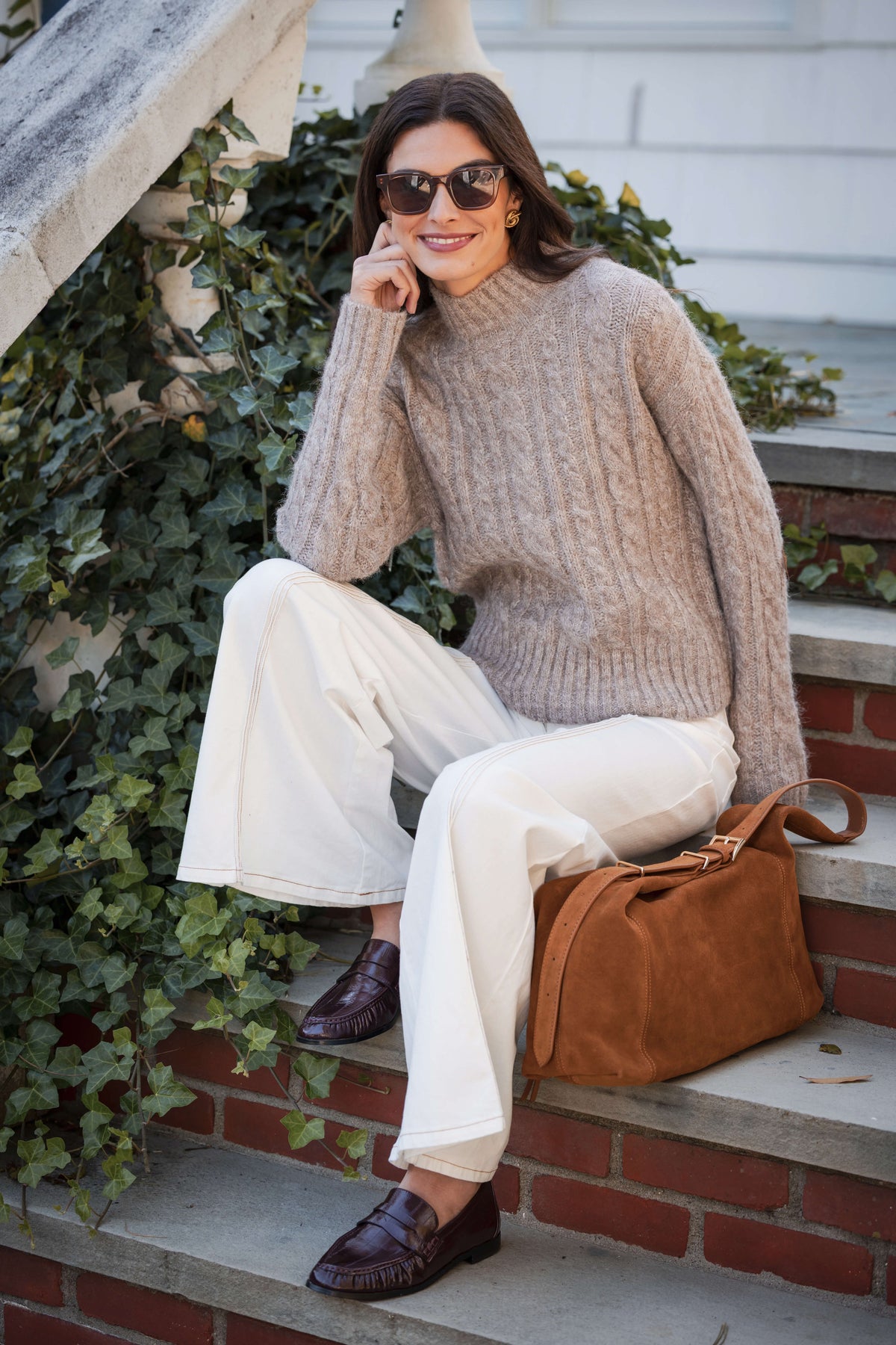 Beige cable knit sweater with crew neckline worn with white pants and brown leather bag.