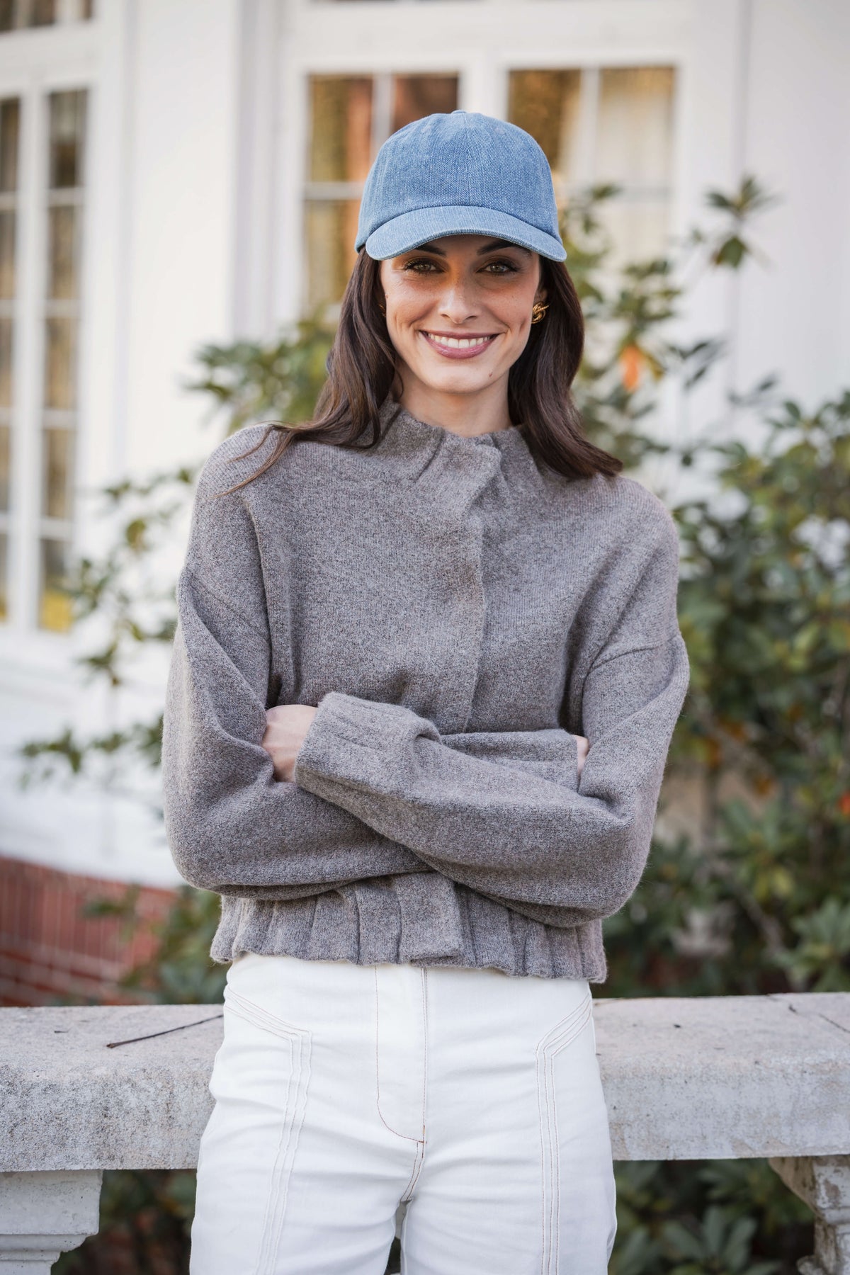 Gray knit sweater with pleated ruffle hem detail at waist, worn with white pants and light blue denim cap.