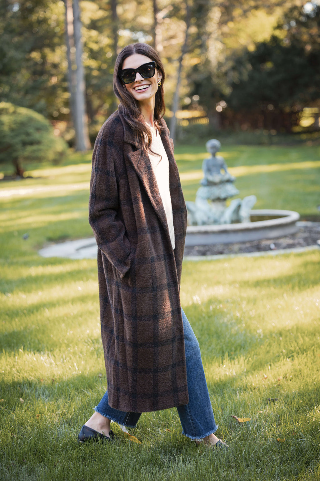 Woman wearing a brown plaid cocoon coat over a white shirt and blue jeans, outdoors.