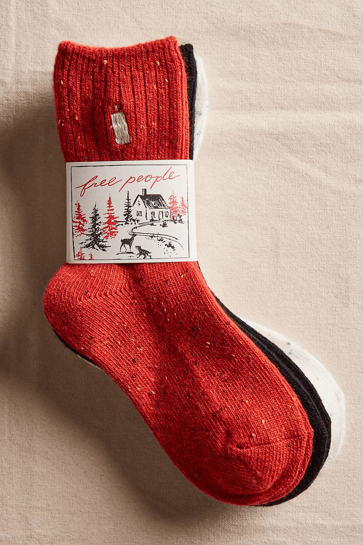 Three stacked flecked knit socks in red, black, and cream are bundled with a paper band showing a snowy cabin scene.