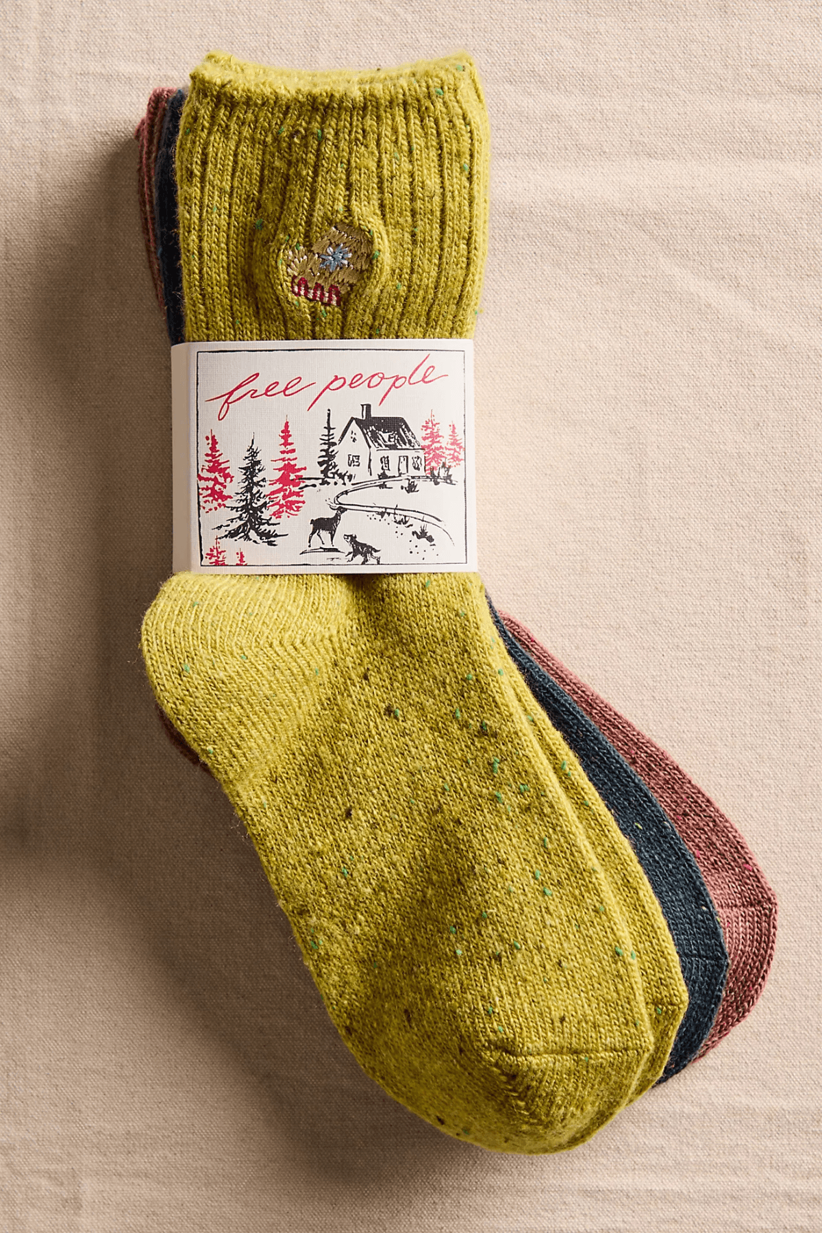 Three pairs of speckled knit socks in chartreuse, navy and rust are bundled with a paper band showing a snowy cabin illustration.
