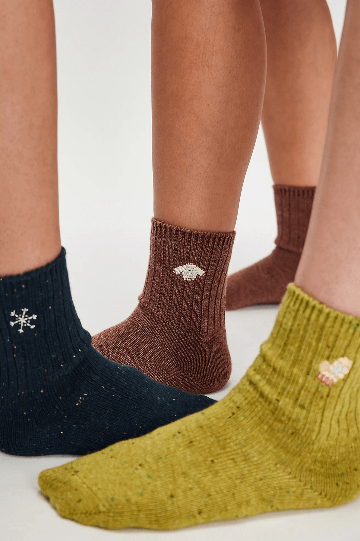 Three people's feet wear ribbed ankle socks in navy, brown, and chartreuse, each embroidered with a small snowflake, sweater, or mitten.
