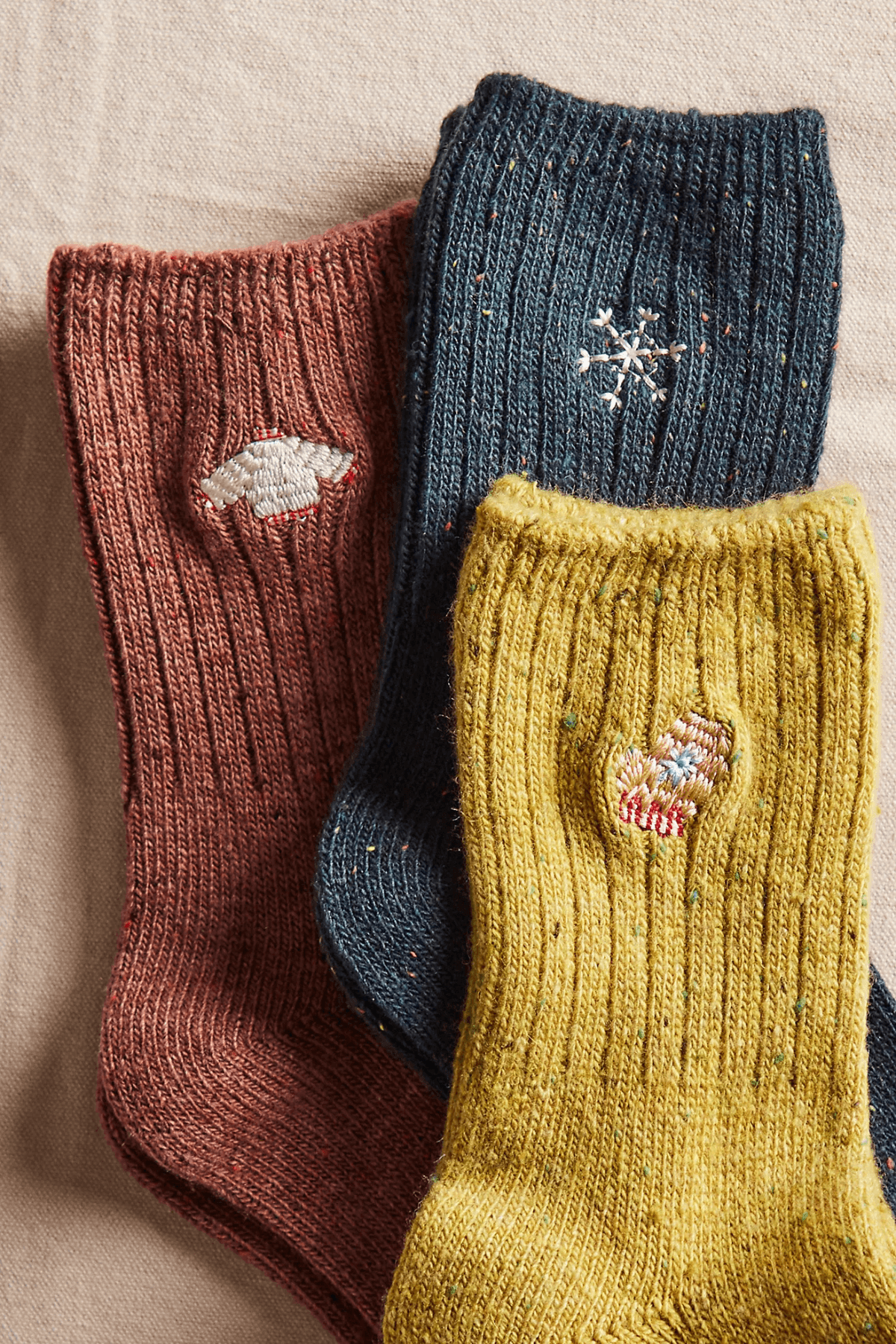 Three ribbed crew socks in rust, dark teal, and mustard yellow, each with a small embroidered winter motif, lie on linen.