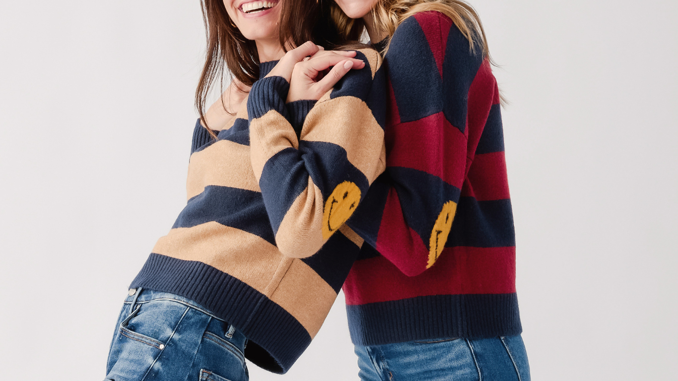 Two women wearing striped knit sweaters with yellow smiley face details, posed against a light gray studio background.