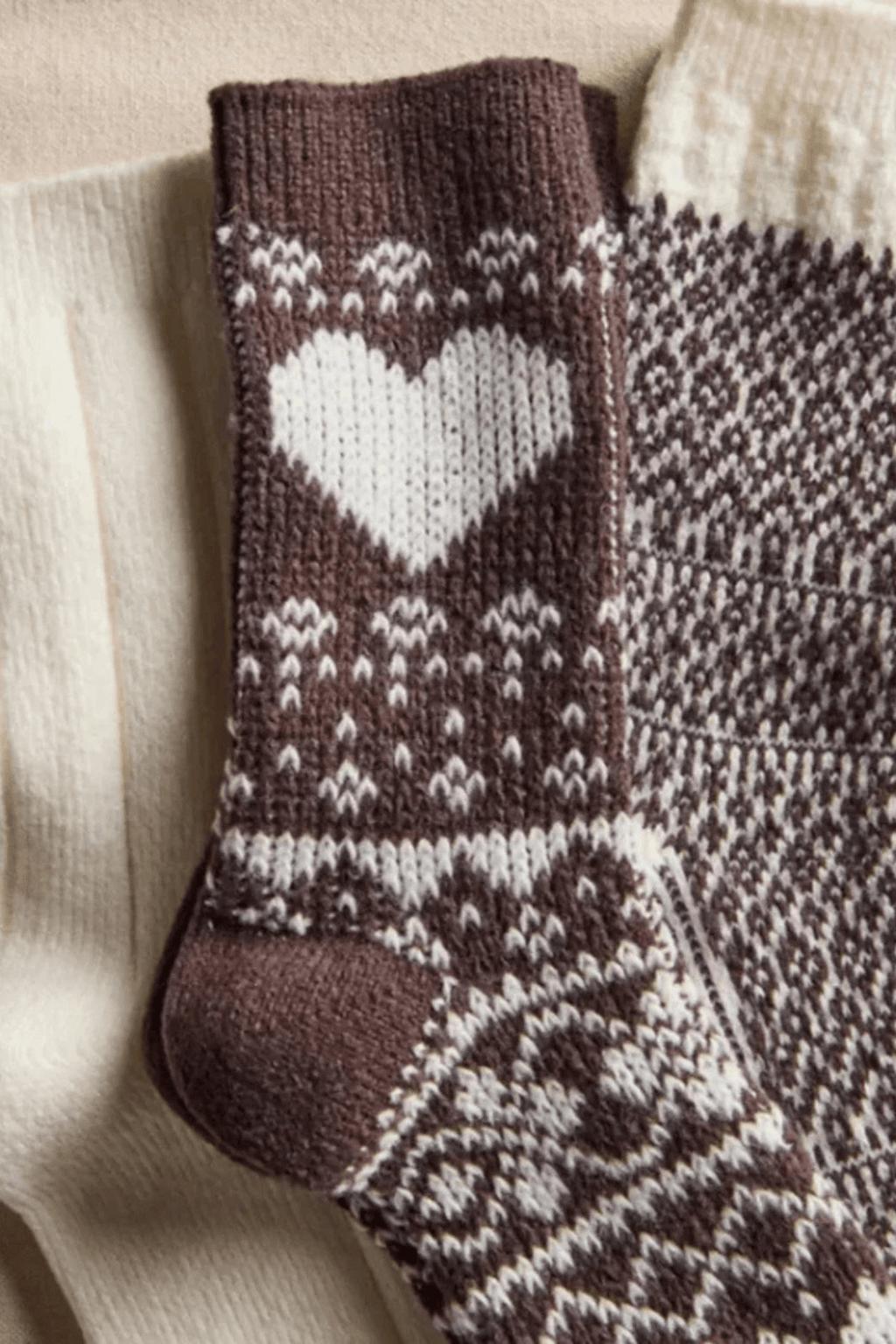 Brown knit socks with a white heart and Nordic patterns lie beside solid cream socks on a beige surface.