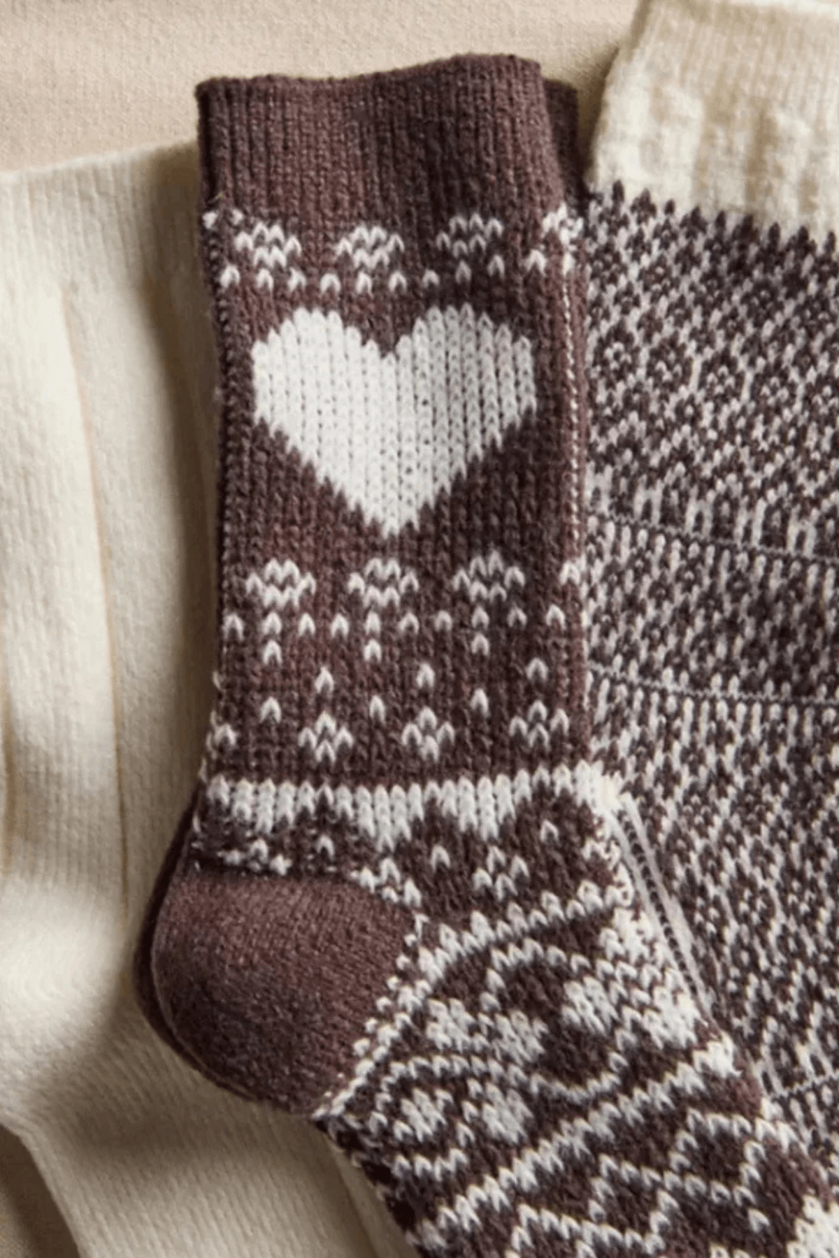 Brown knit socks with a white heart and Nordic patterns lie beside solid cream socks on a beige surface.