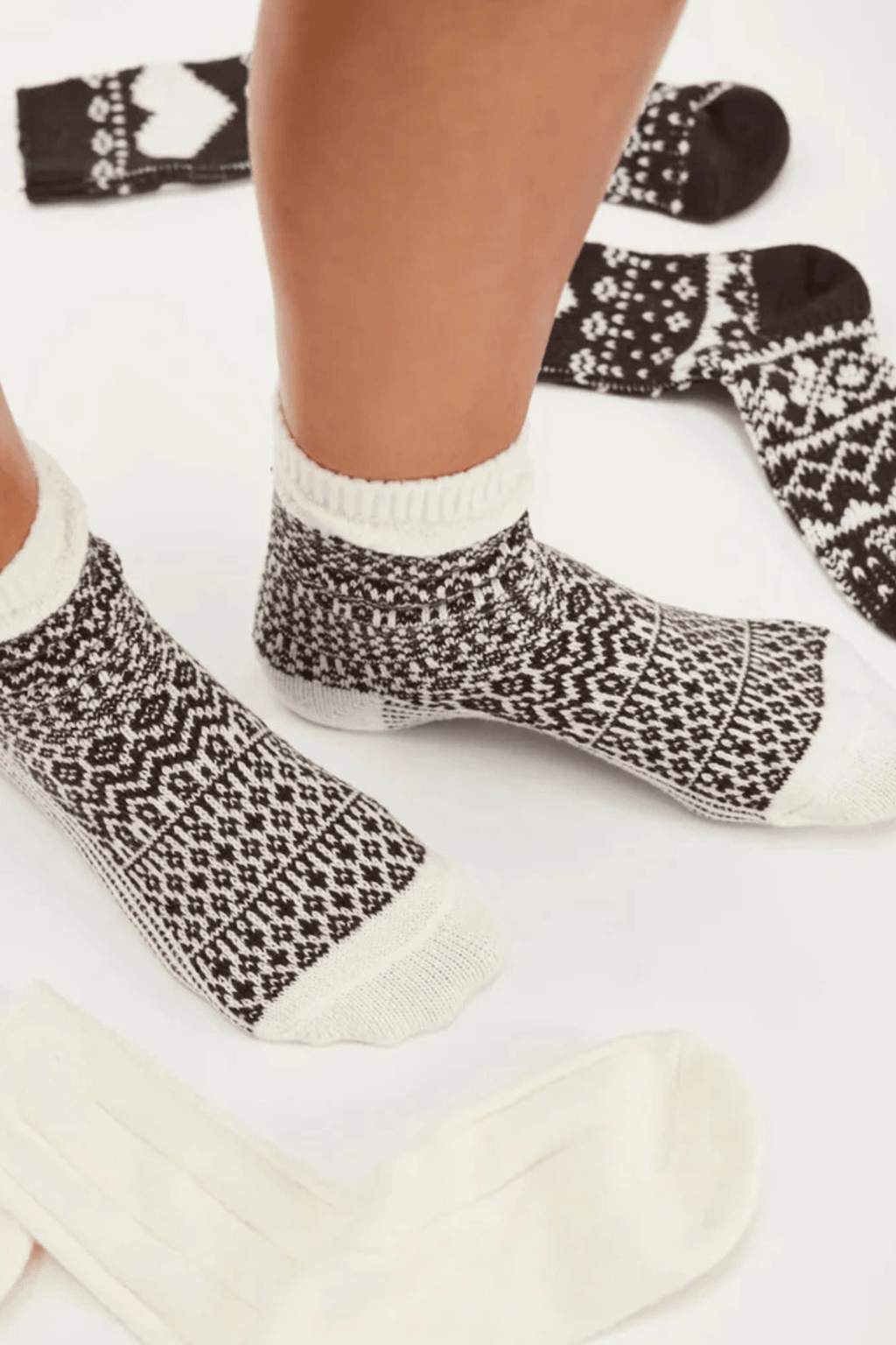 Two feet wear brown and cream patterned knit ankle socks, with more dark Nordic-print socks and plain cream socks laid nearby.