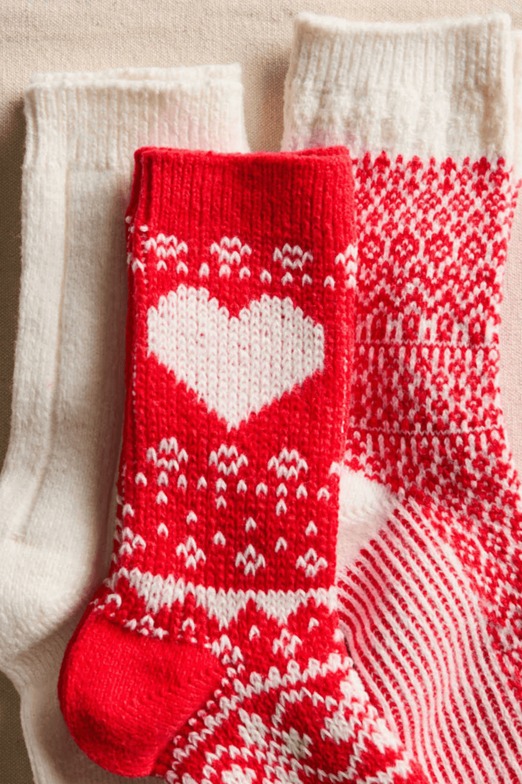 Three knit socks lie together: a red one with a white heart, a red and white patterned one, and a cream one.