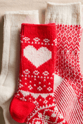 Three knit socks lie together: a red one with a white heart, a red and white patterned one, and a cream one.