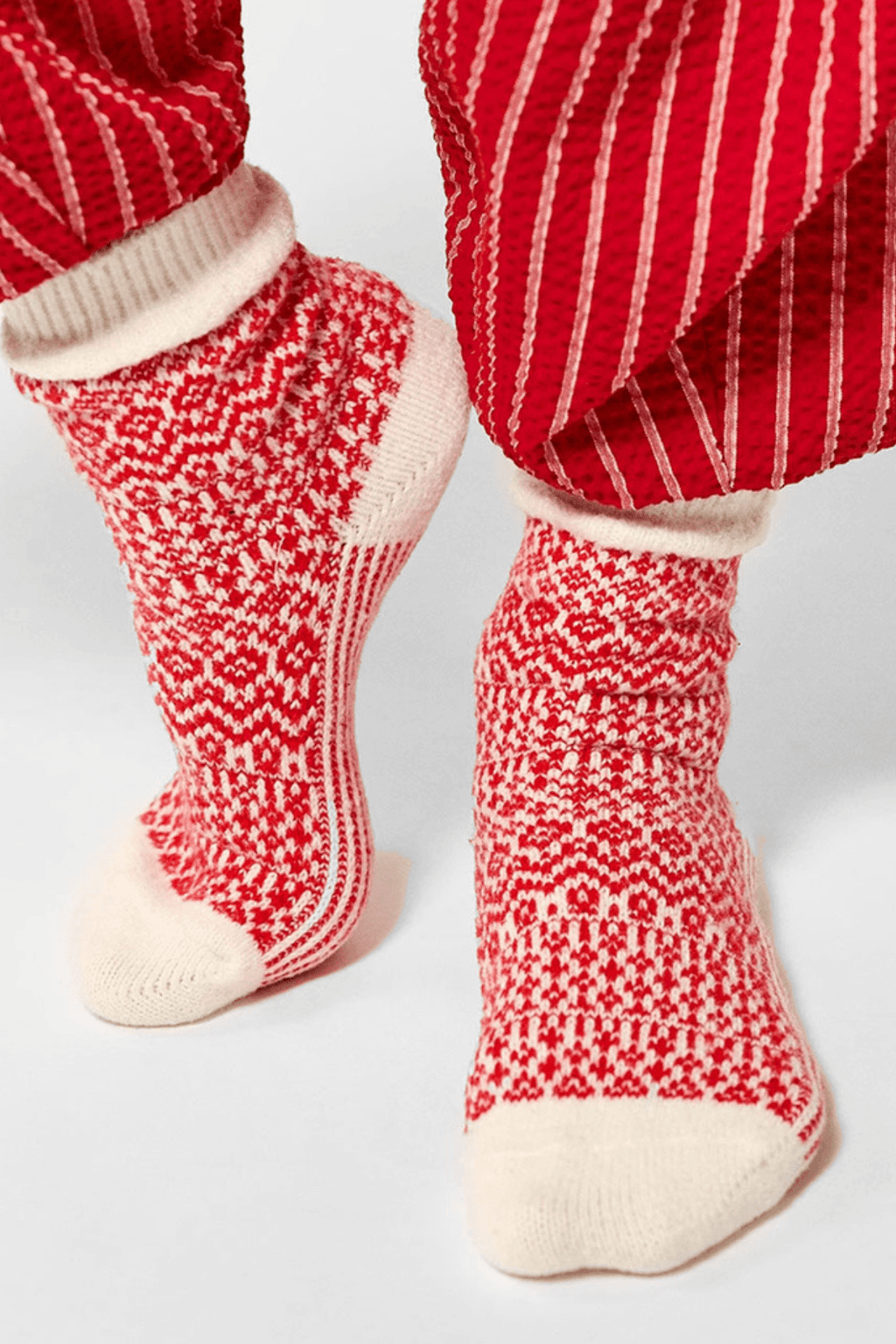 Red and cream patterned knit socks with cream heels and toes are worn beneath red pinstriped pants against a plain light background.