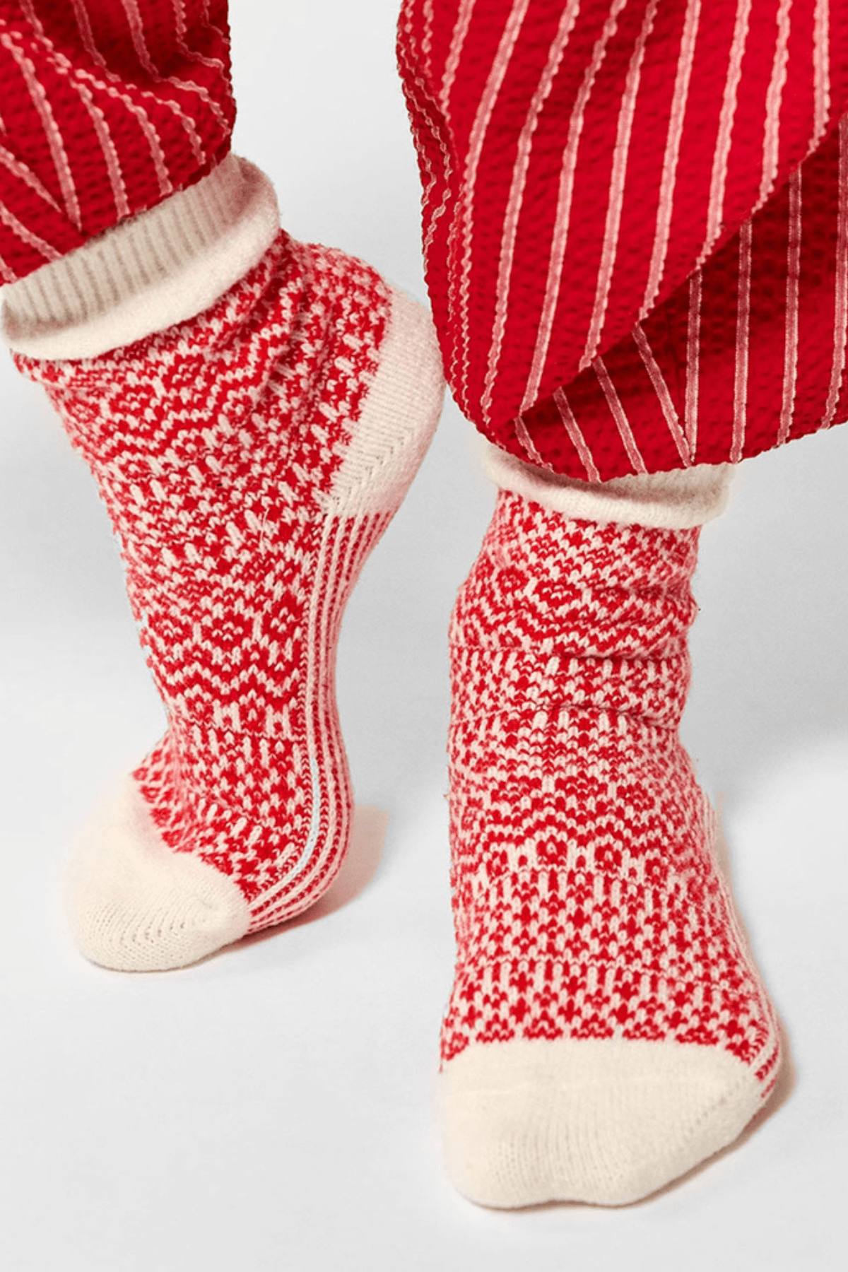 Red and cream patterned knit socks with cream heels and toes are worn beneath red pinstriped pants against a plain light background.