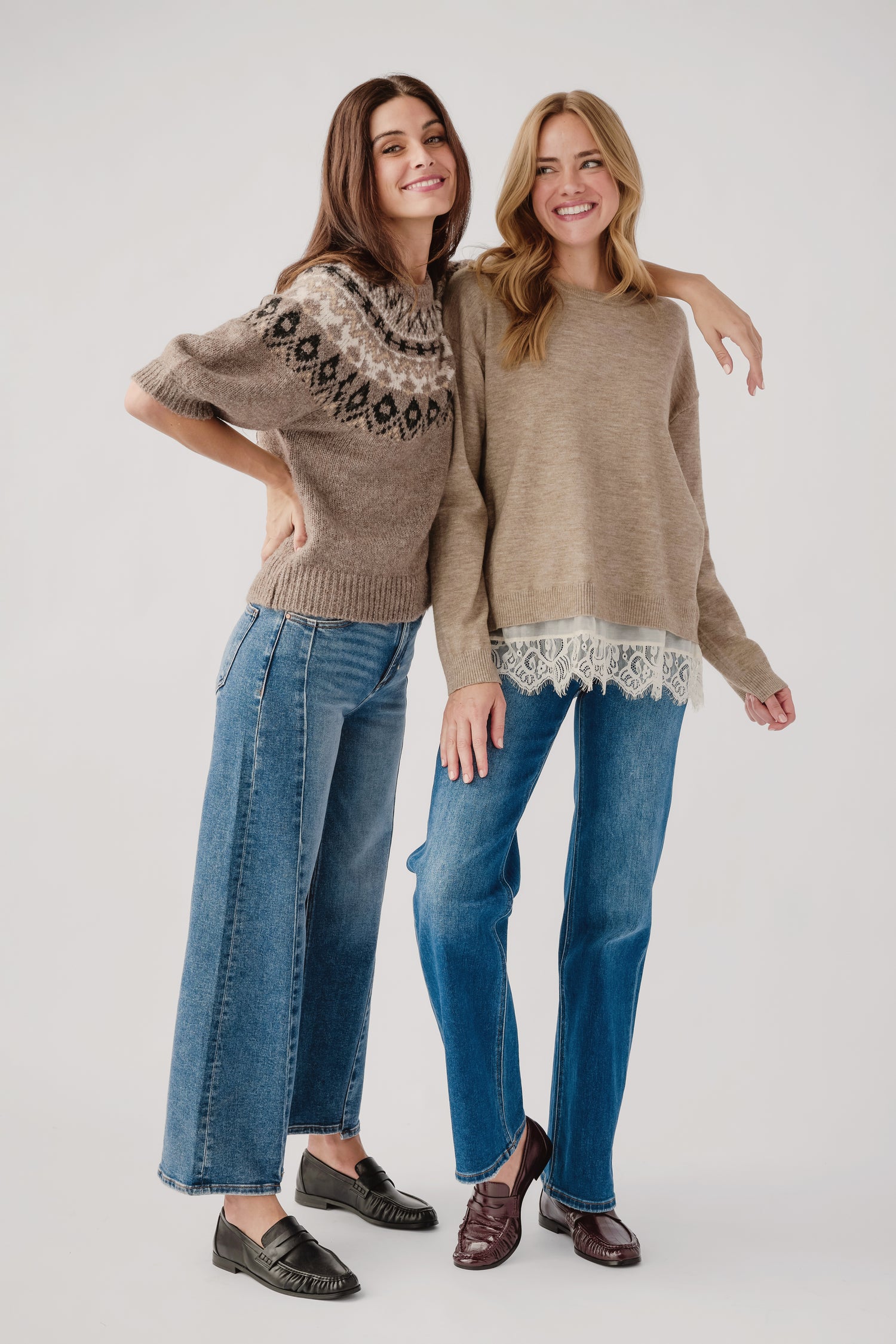 Two women modeling cozy knit sweaters and wide-leg blue jeans against a light gray studio background. The woman on the left wears a taupe knit sweater with a dark geometric pattern across the chest and sleeves, paired with dark loafers.