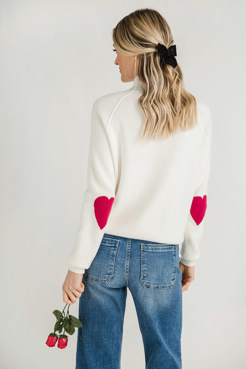 White sweater with red heart patches on sleeves and ribbed high collar, back view