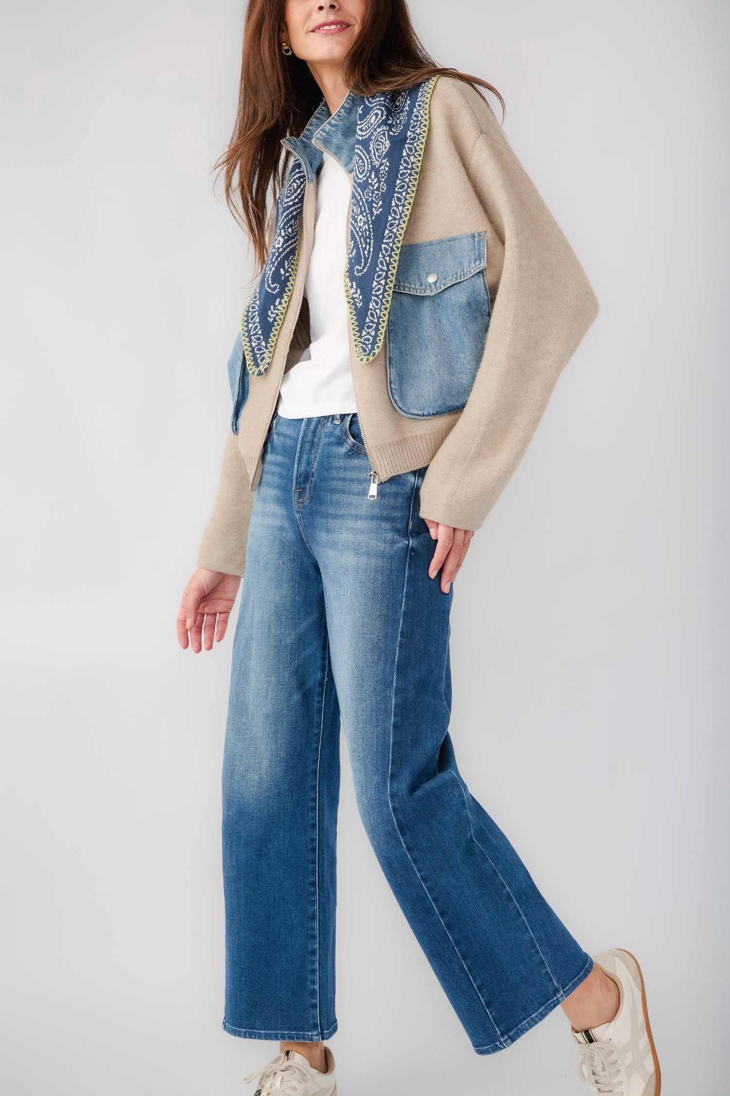 A woman models a beige knit cardigan with blue denim pockets and a blue paisley scarf detail on the collar, paired with blue jeans against a neutral gray background.