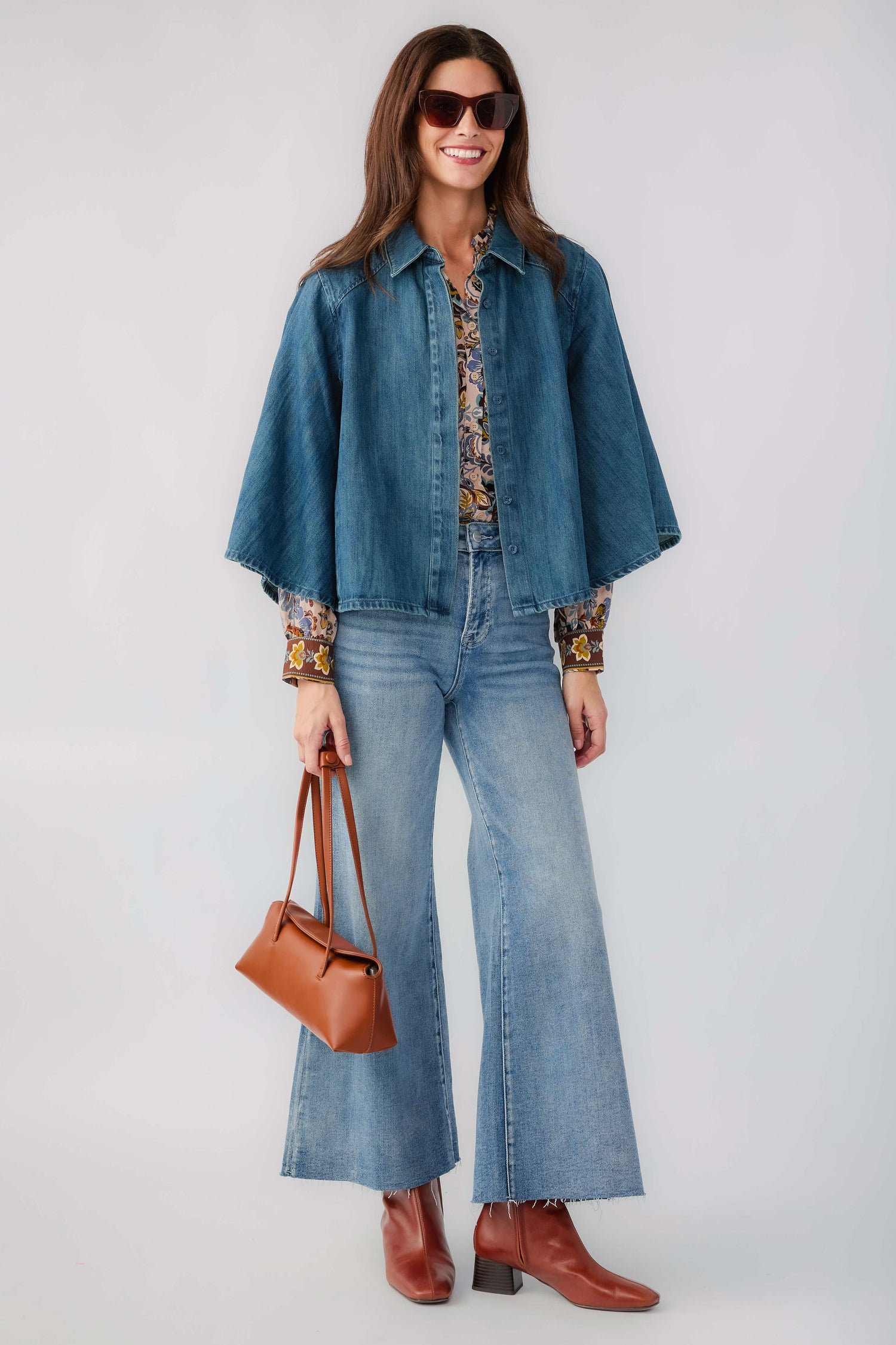 A woman modeling a blue denim shirt-jacket and matching wide-leg jeans, styled with a patterned blouse, brown leather handbag, and brown ankle boots against a light gray studio background.