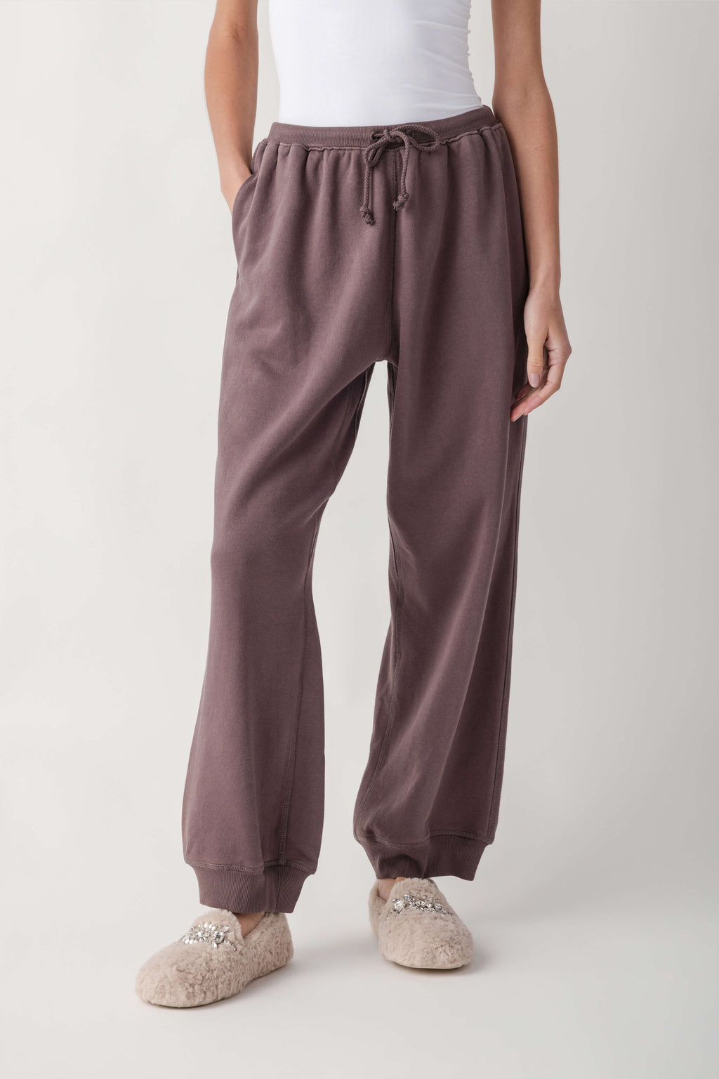 A person wearing loose, wide-leg jogger-style lounge pants in a muted dusty mauve/taupe color, paired with a white top and fuzzy slip-on slippers, photographed against a clean light gray studio background.