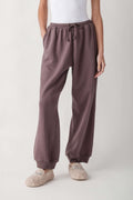 A person wearing loose, wide-leg jogger-style lounge pants in a muted dusty mauve/taupe color, paired with a white top and fuzzy slip-on slippers, photographed against a clean light gray studio background.
