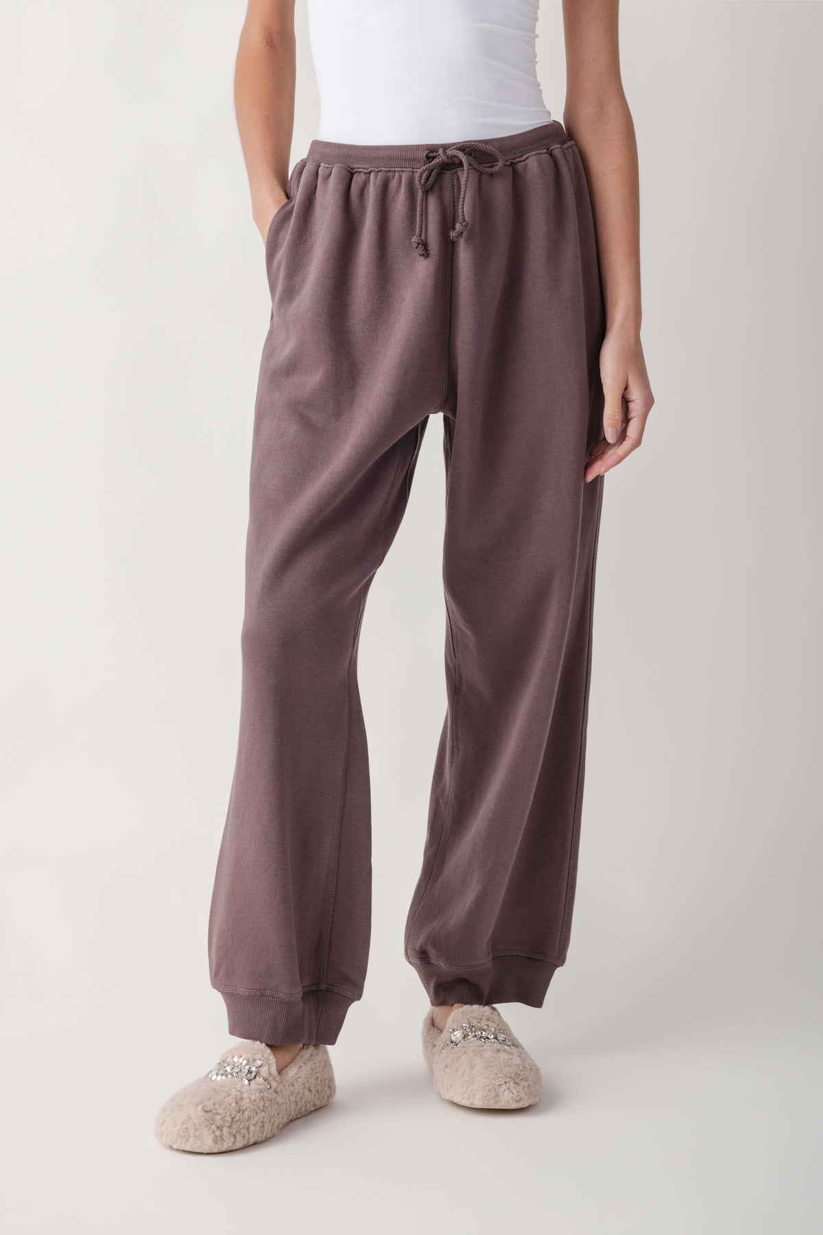 A person wearing loose, wide-leg jogger-style lounge pants in a muted dusty mauve/taupe color, paired with a white top and fuzzy slip-on slippers, photographed against a clean light gray studio background.