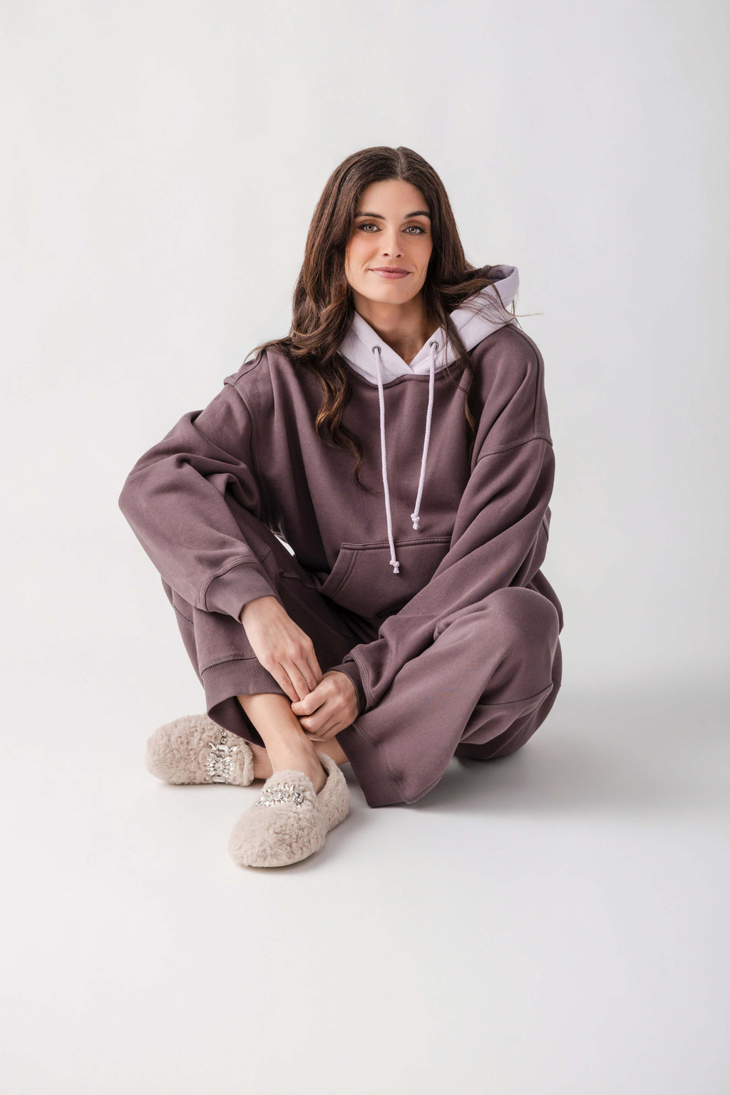 A woman sits cross-legged on a light gray studio floor wearing an oversized mauve-brown hoodie and matching sweatpants with a light gray inner hood lining, white drawstrings, and fuzzy beige slip-on slippers.