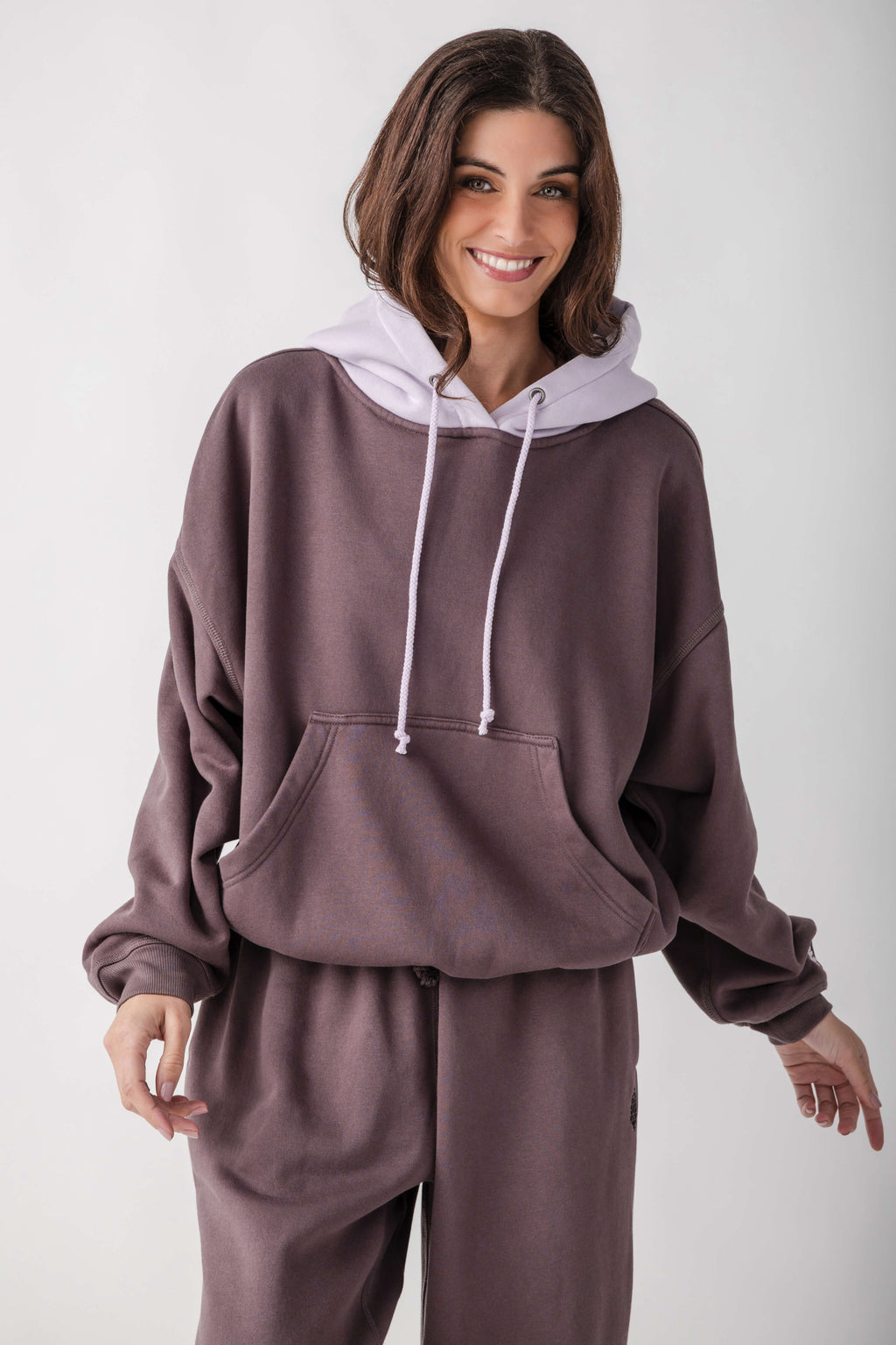 A woman wearing a matching two-piece loungewear set in a dusty mauve-brown color, featuring a pullover hoodie with a contrasting light lavender hood and matching sweatpants, shot against a plain light gray background.