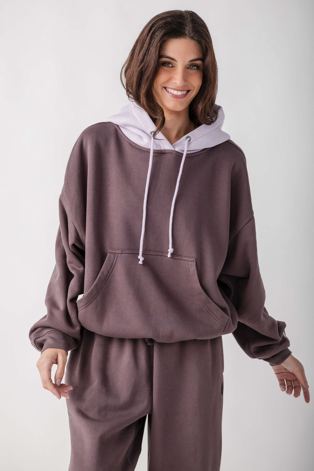 A woman wearing a matching two-piece loungewear set in a dusty mauve-brown color, featuring a pullover hoodie with a contrasting light lavender hood and matching sweatpants, shot against a plain light gray background.