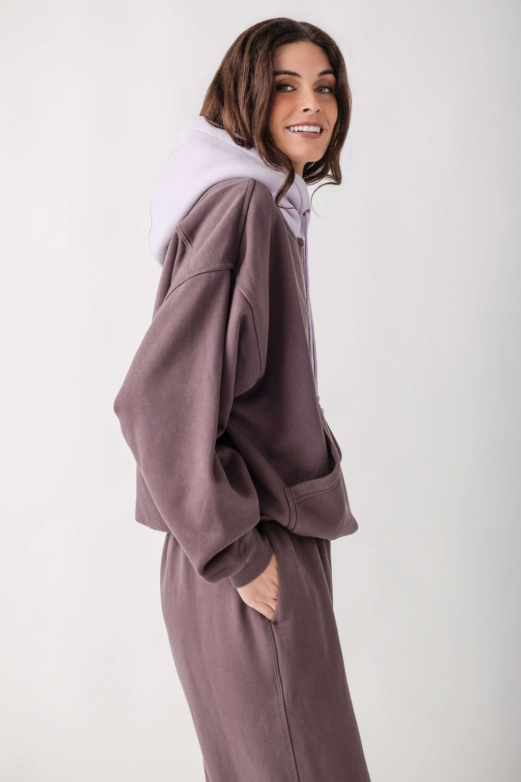 A woman wearing a matching lounge set consisting of an oversized zip-up sweatshirt and sweatpants in a dusty mauve-brown color, layered with a light lavender hoodie, against a plain light gray studio background.
