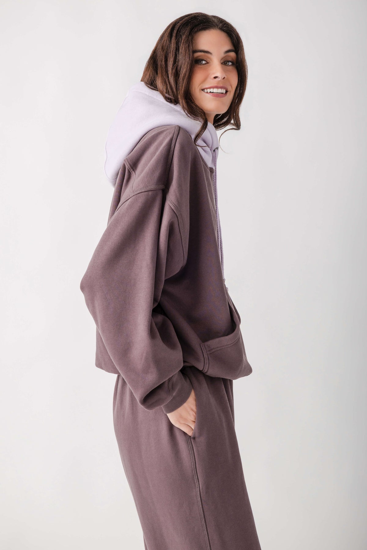 A woman wearing a matching lounge set consisting of an oversized zip-up sweatshirt and sweatpants in a dusty mauve-brown color, layered with a light lavender hoodie, against a plain light gray studio background.
