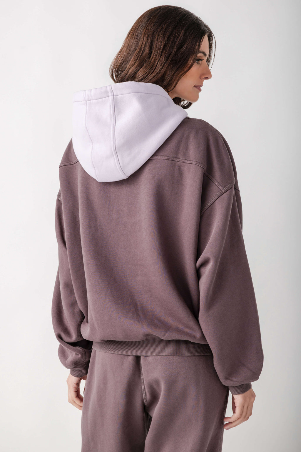 A woman shown from the back wearing a dusty mauve sweatshirt with a contrasting light lavender hood and matching sweatpants against a plain light gray background.