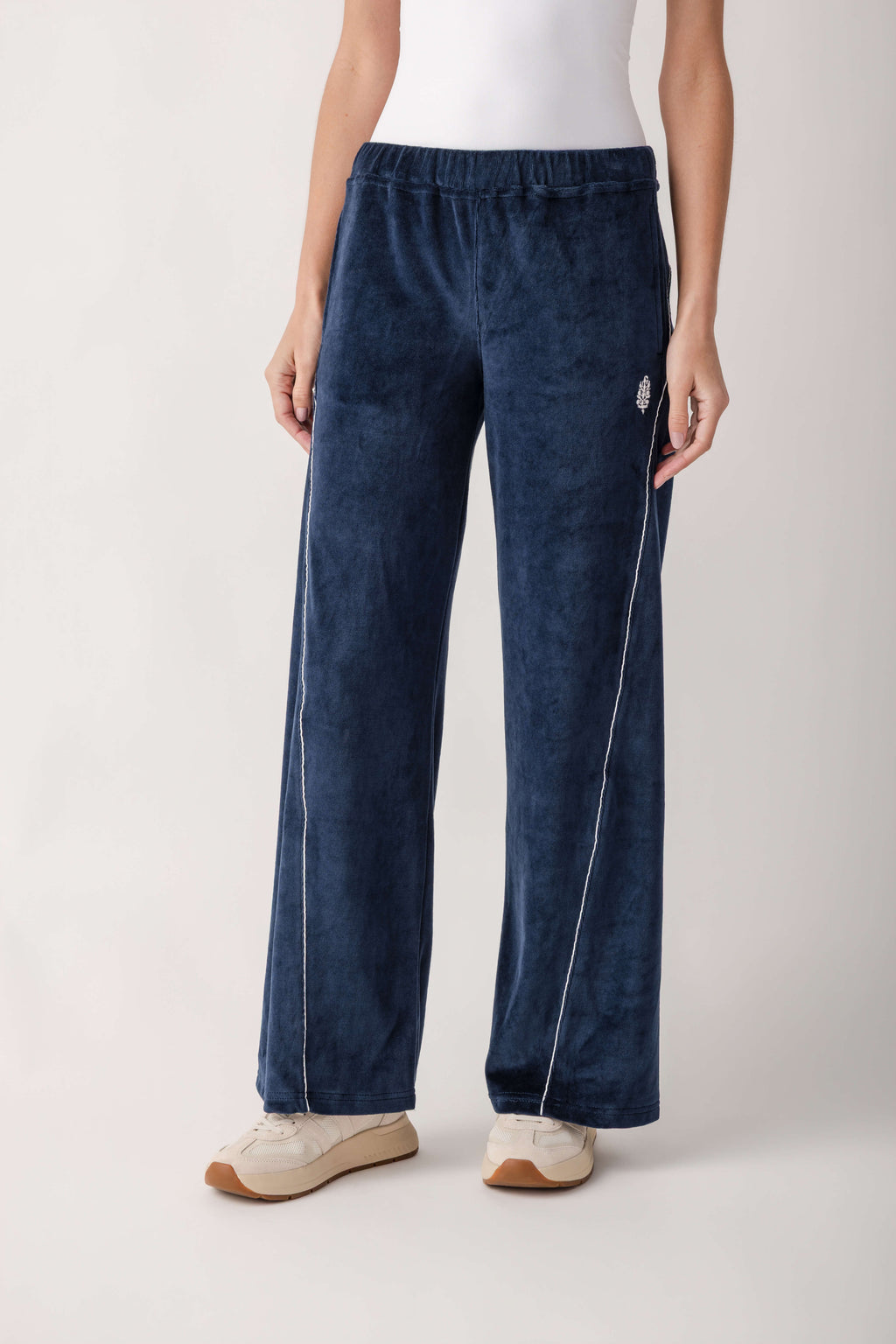 A person wears navy blue velour wide-leg pants with white piping, an elastic waistband, a small embroidered logo, and cream sneakers.