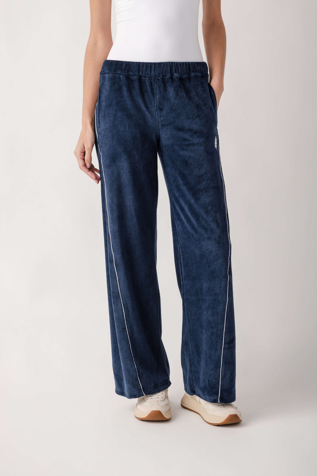 A person wears navy blue velour wide-leg pants with white piping along the sides, a white top, and cream sneakers.