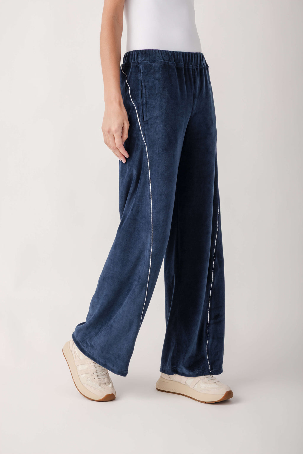A person wears wide-leg navy velour pants with white piping along the sides, paired with a white top and cream sneakers.