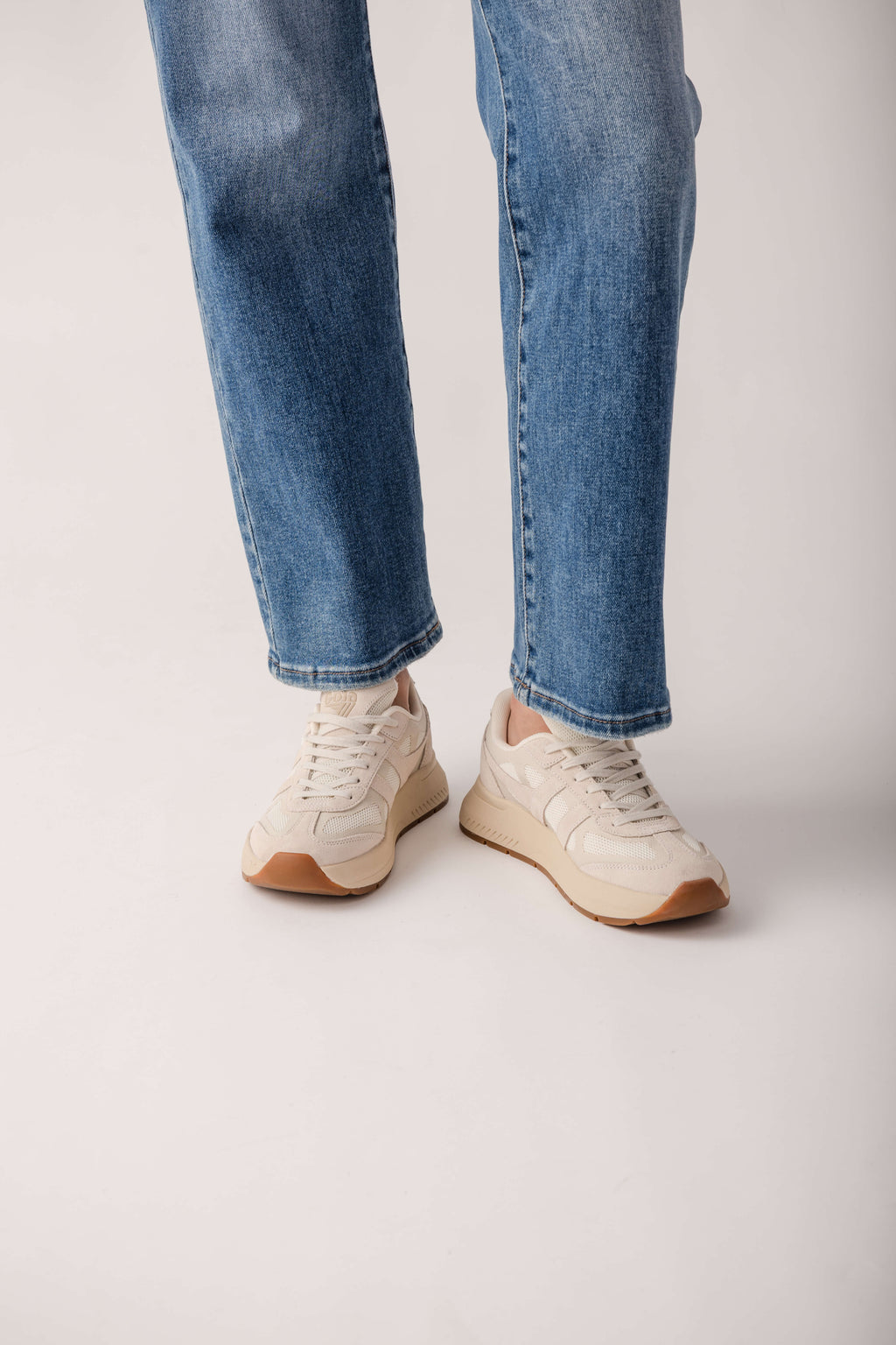 Cream lace-up sneakers with tan sole worn with blue jeans.