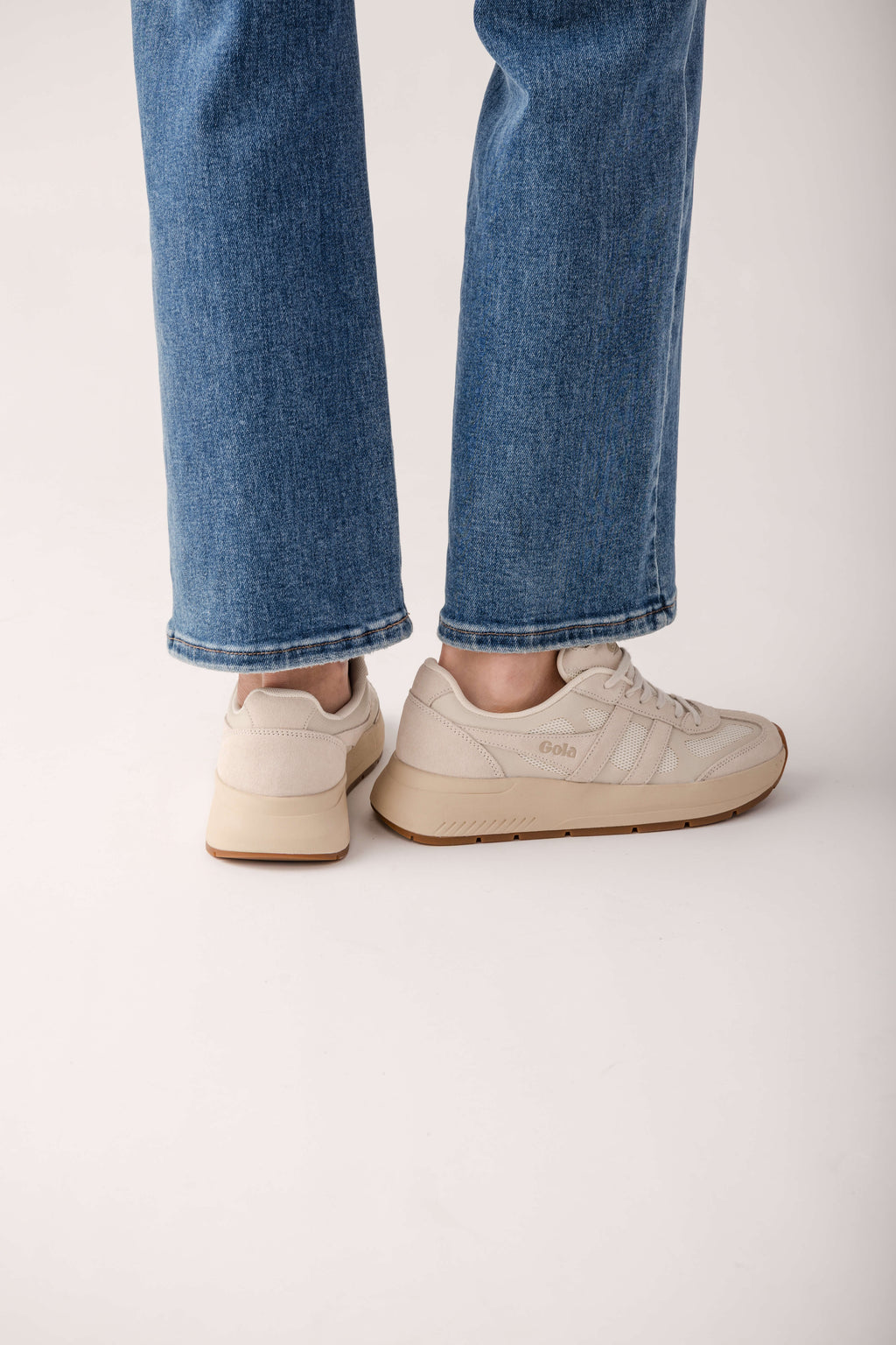 Cream-colored lace-up sneakers with textured upper and gum sole, worn with blue jeans.