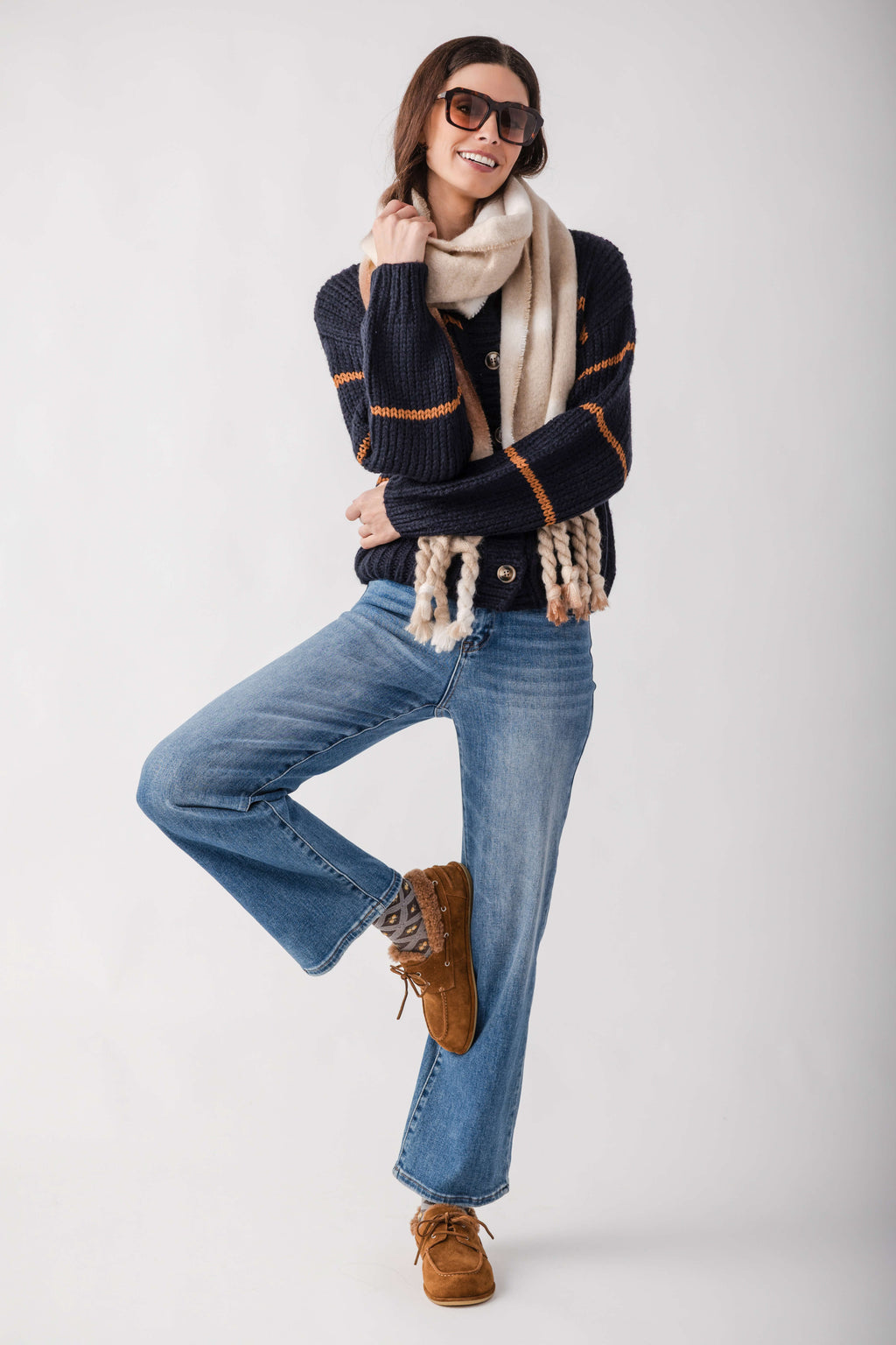 A woman modeling a dark navy knit sweater with orange stripes, a cream scarf, blue jeans, brown suede boots, and sunglasses against a light gray studio background.