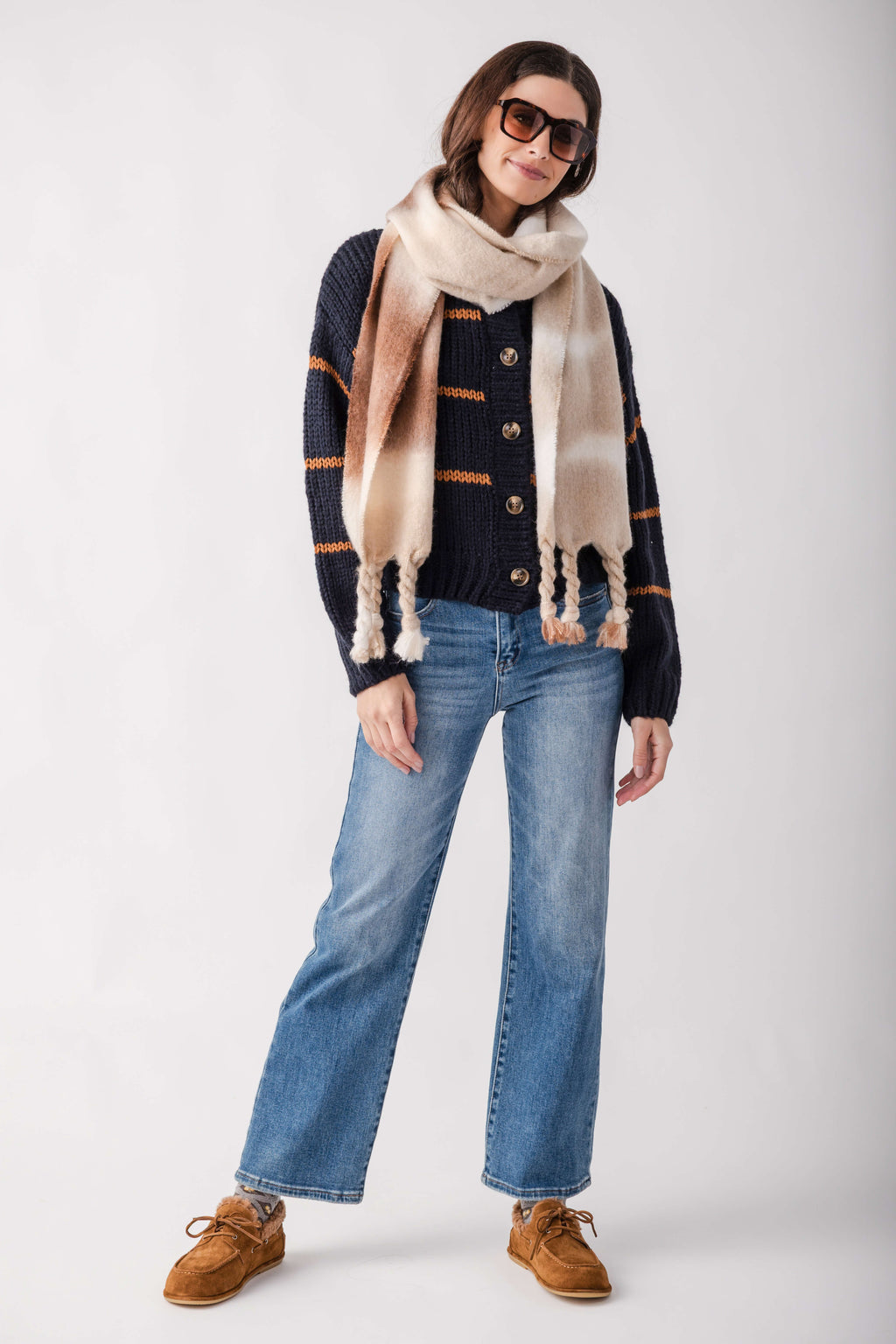 A woman modeling a dark navy knit cardigan with orange stripes, a beige and cream striped scarf, blue jeans, and brown suede shoes against a light gray studio background.