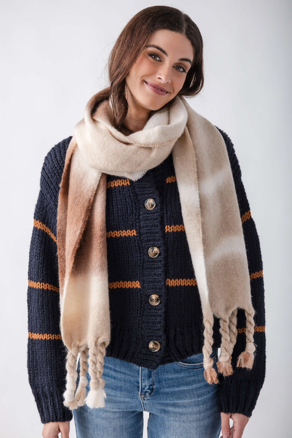 Cream and tan blanket scarf with frayed edges worn draped around shoulders over navy and orange striped cardigan.