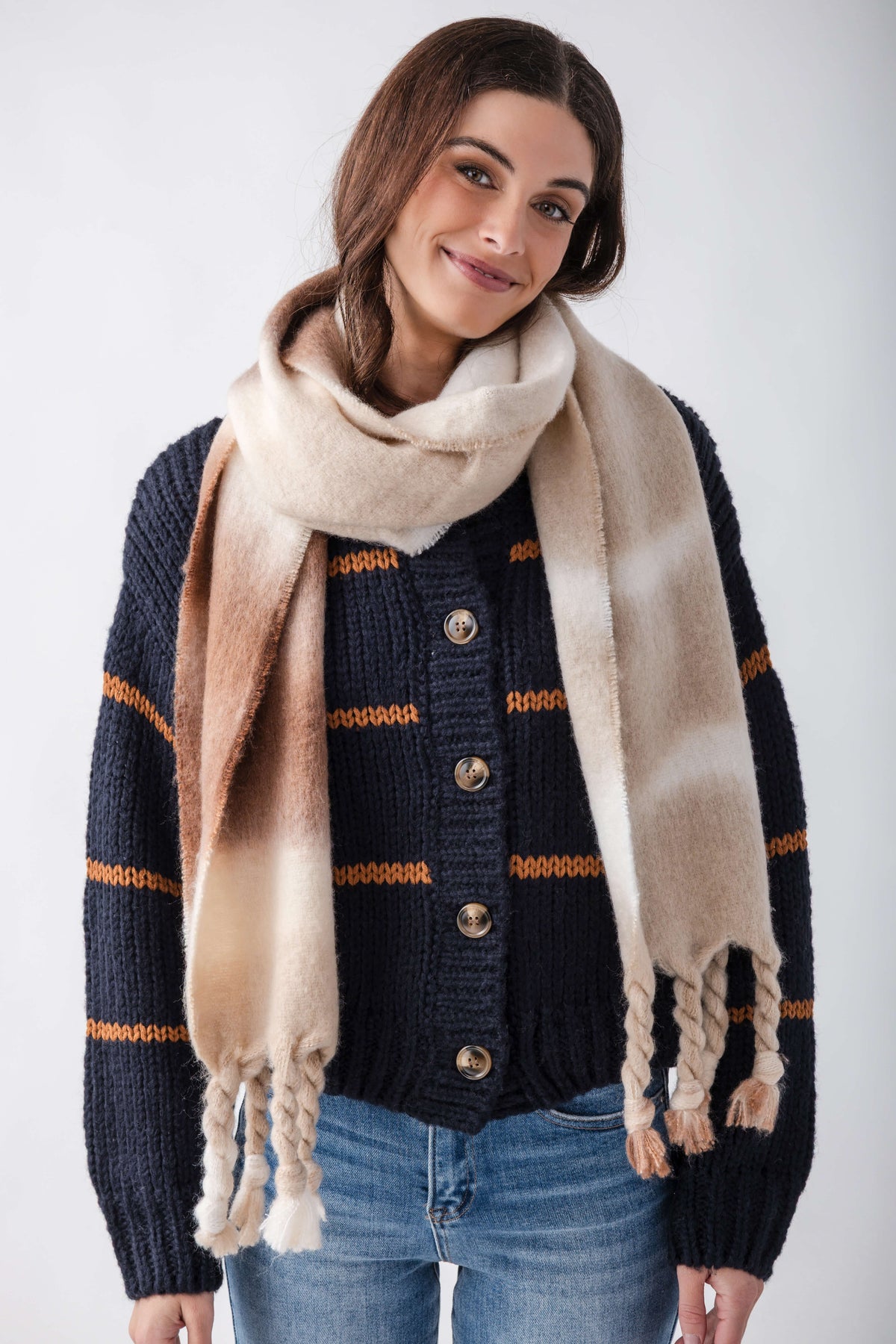 Cream and tan blanket scarf with frayed edges worn draped around shoulders over navy and orange striped cardigan.