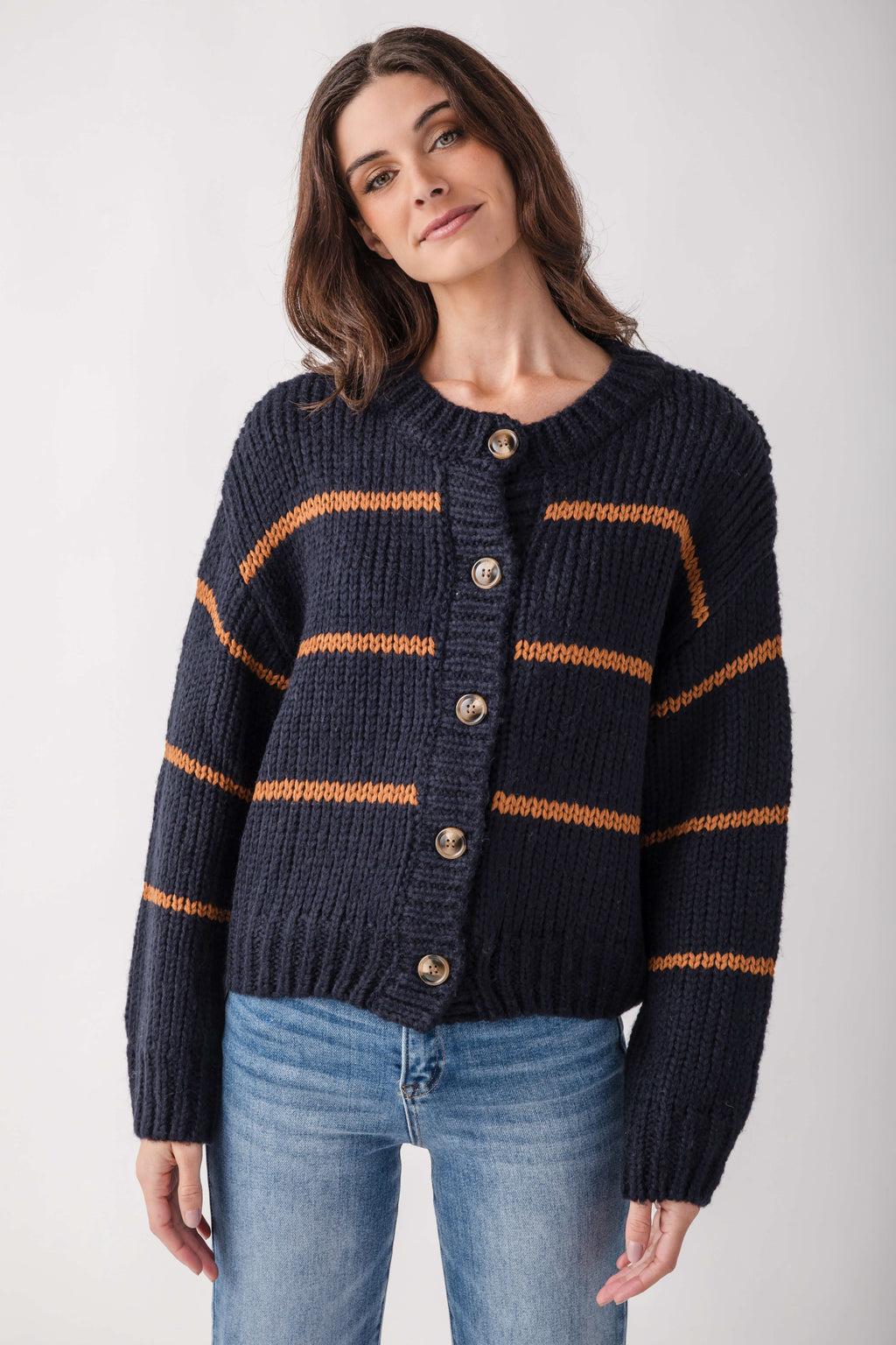 A woman wearing a dark navy blue chunky knit cardigan with horizontal rust-orange stripes and brown buttons, paired with blue jeans, against a light gray studio background.