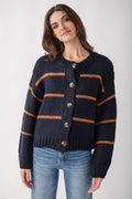 A woman wearing a dark navy blue chunky knit cardigan with horizontal rust-orange stripes and brown buttons, paired with blue jeans, against a light gray studio background.