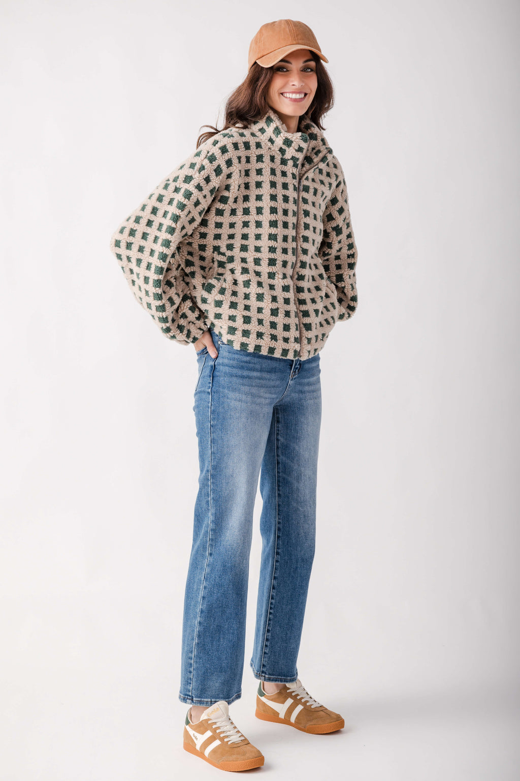 A woman modeling a beige and green checkered zip-up jacket, blue jeans, and tan sneakers against a plain light gray studio background.