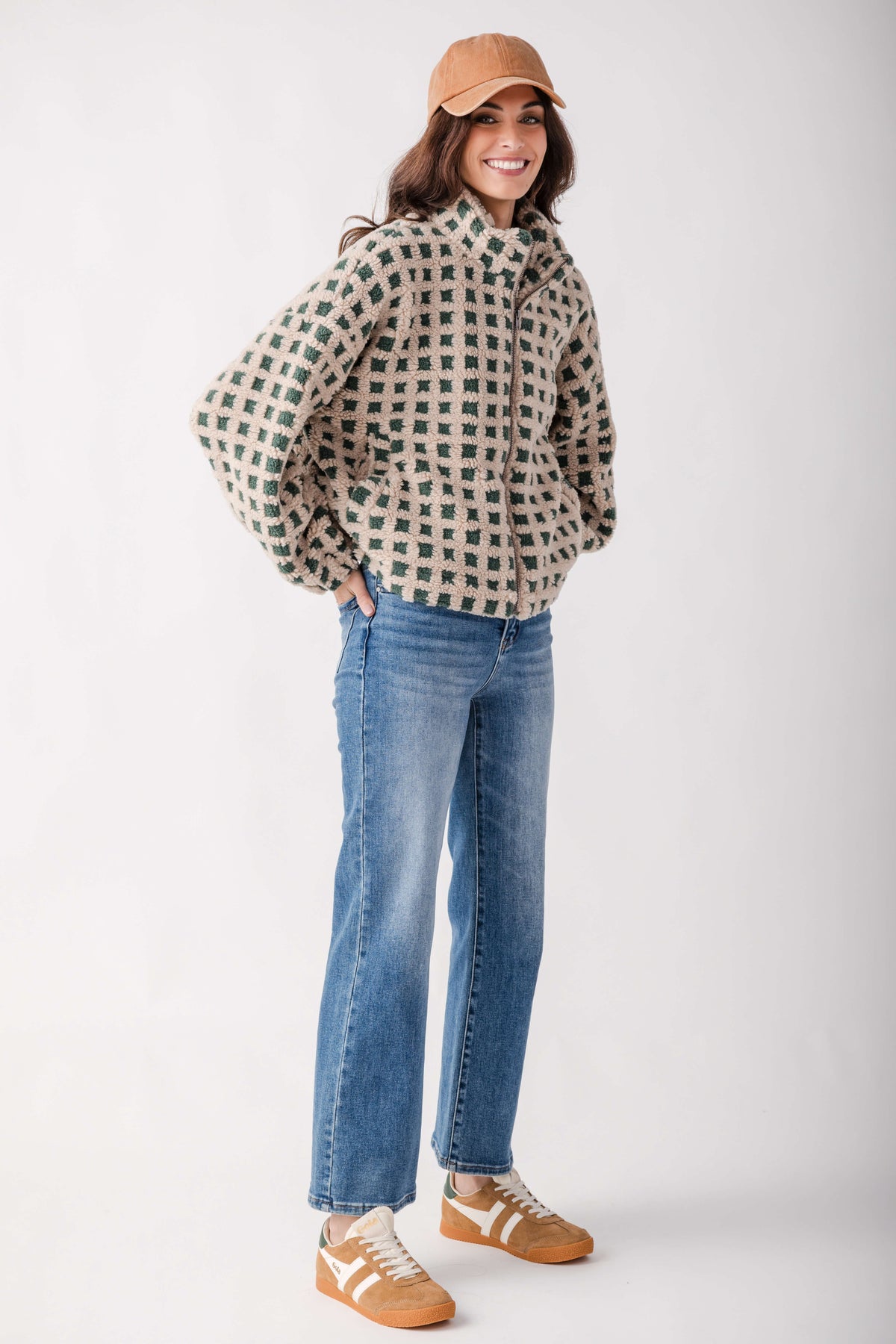 A woman modeling a beige and green checkered zip-up jacket, blue jeans, and tan sneakers against a plain light gray studio background.