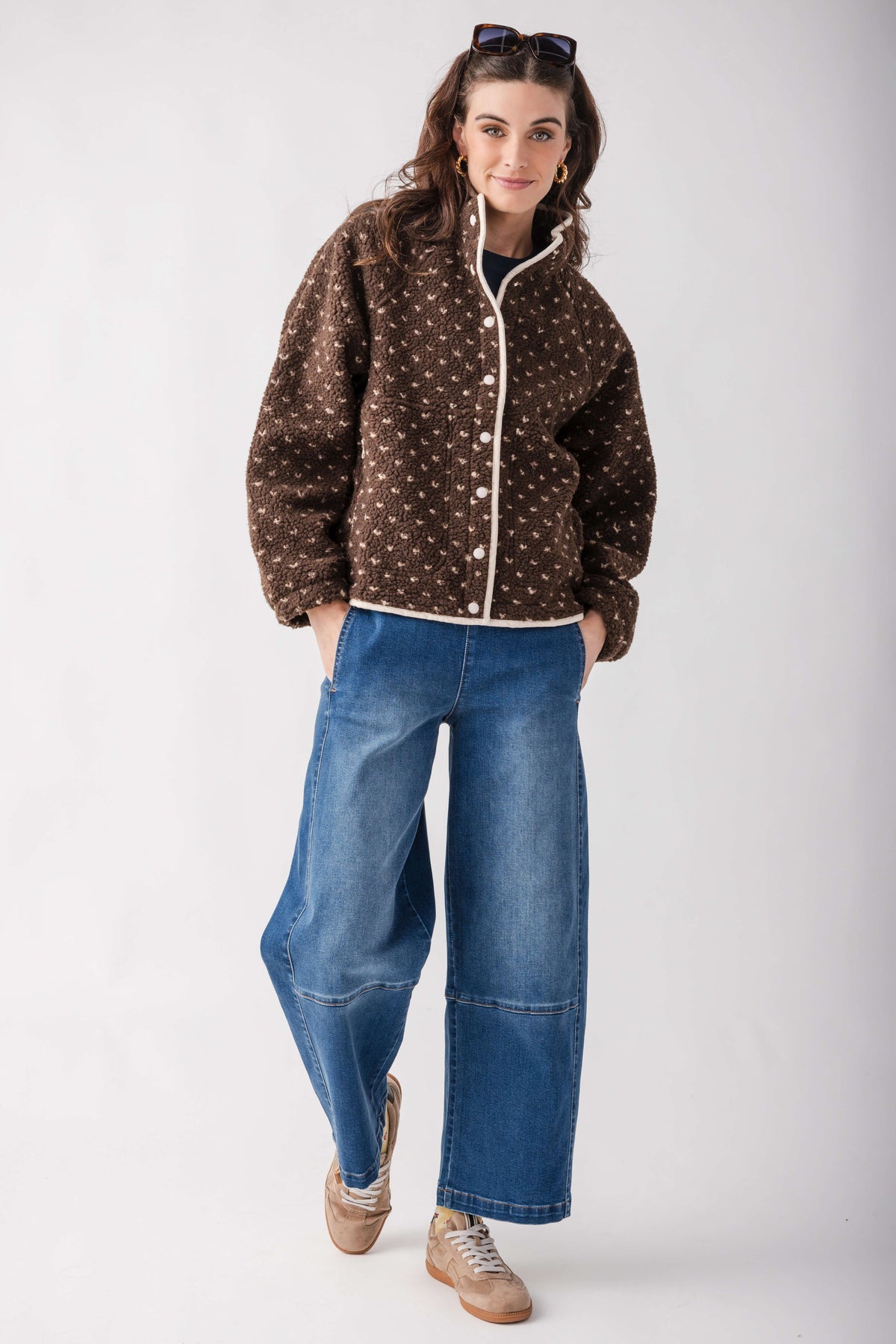 A woman modeling a brown button-up fleece jacket with a white polka dot pattern, paired with wide-leg blue jeans and beige sneakers, against a plain light gray studio background.