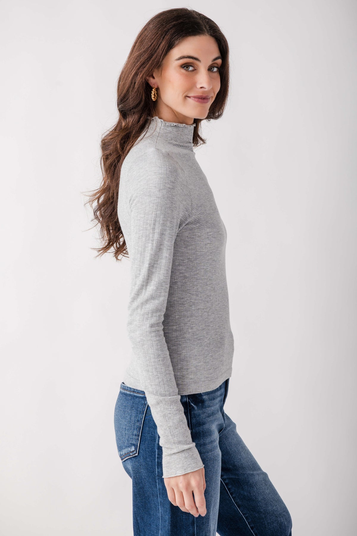 Woman wearing light gray ribbed mock neck long-sleeve top with blue jeans, shown from the side.
