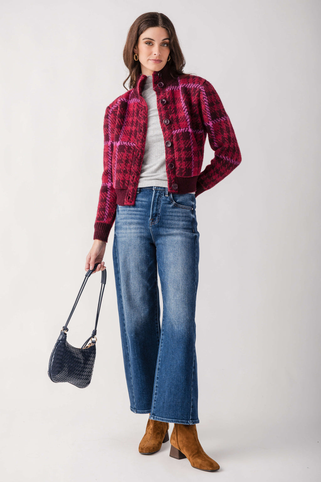 A woman models a red and black plaid cropped button-up cardigan over a white t-shirt, paired with wide-leg blue jeans, brown suede boots, and a dark blue handbag, set against a plain light gray studio background.