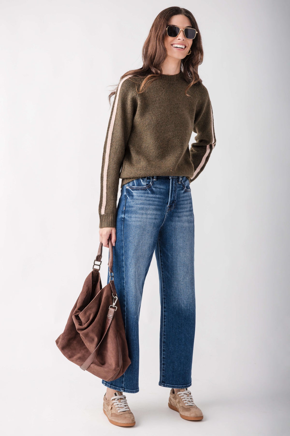 A woman modeling an olive green knit sweater with white stripes down the sleeves, paired with blue jeans, beige sneakers, and holding a brown suede hobo bag against a light gray studio background.