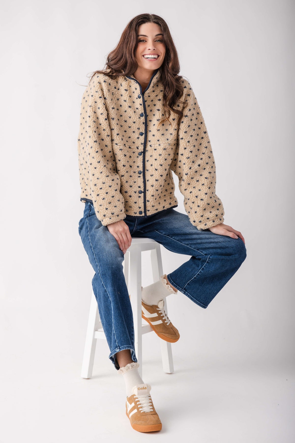 A woman models a beige sherpa jacket with a black polka dot pattern, paired with blue jeans, white socks, and tan sneakers, sitting on a white stool against a light gray background.